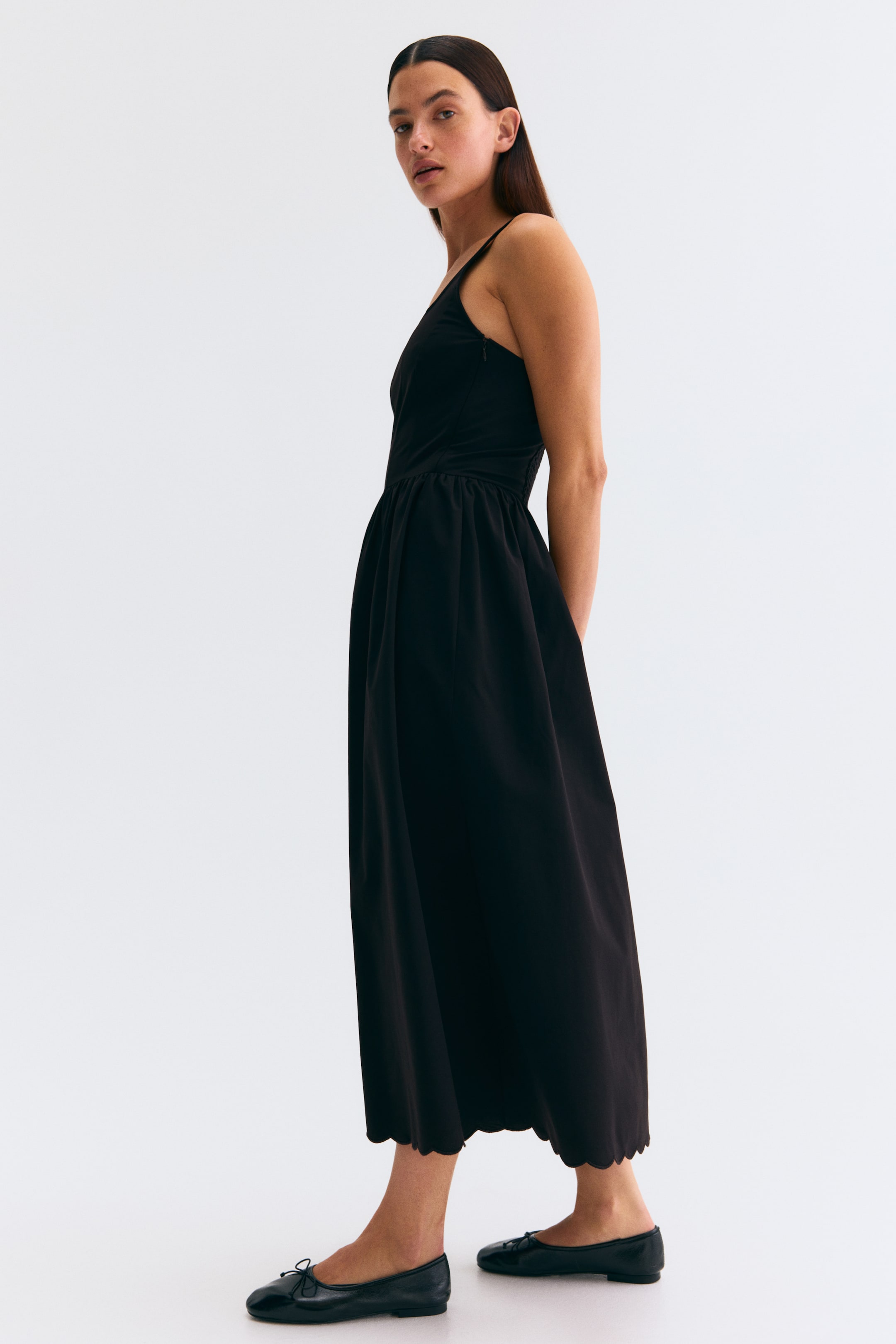 View larger image: A black midi dress is shown in a slightly angled side view, featuring thin spaghetti straps and a fitted bodice. The dress gathers at the waist, flows into a full skirt, and finishes with a scalloped hemline.