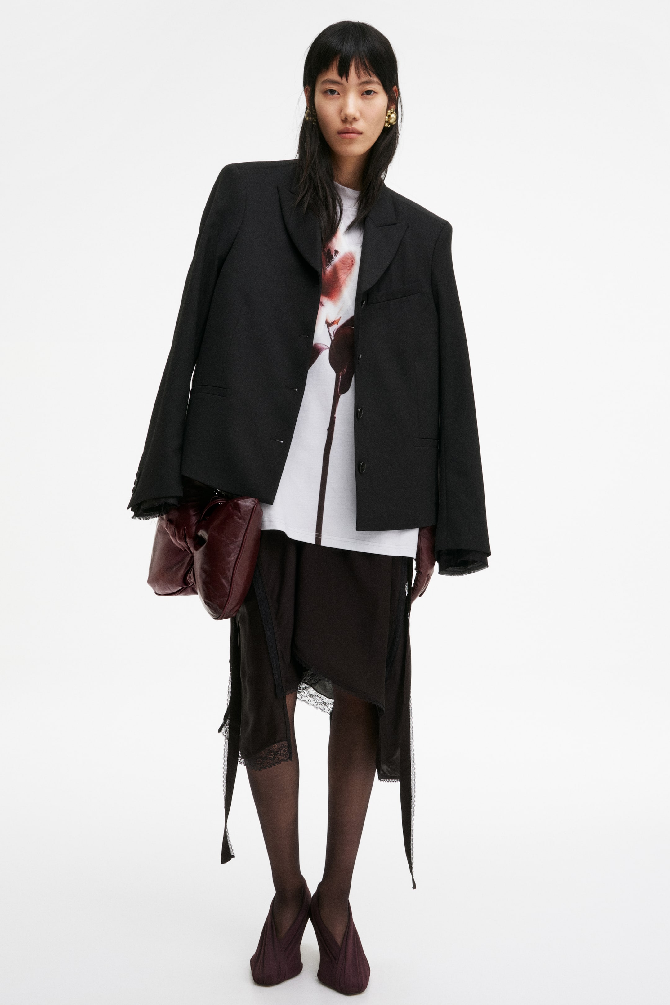 View larger image: The complete ensemble is showcased front-on, featuring an oversized black blazer with prominent shoulder pads and notch lapels, worn open over a white T-shirt adorned with a reddish-brown abstract floral design. It layers an asymmetrical dark brown skirt with lace trim over dark tights, and is accessorised with burgundy long gloves, a ruched pouch handbag, and pointed pleated flat shoes.