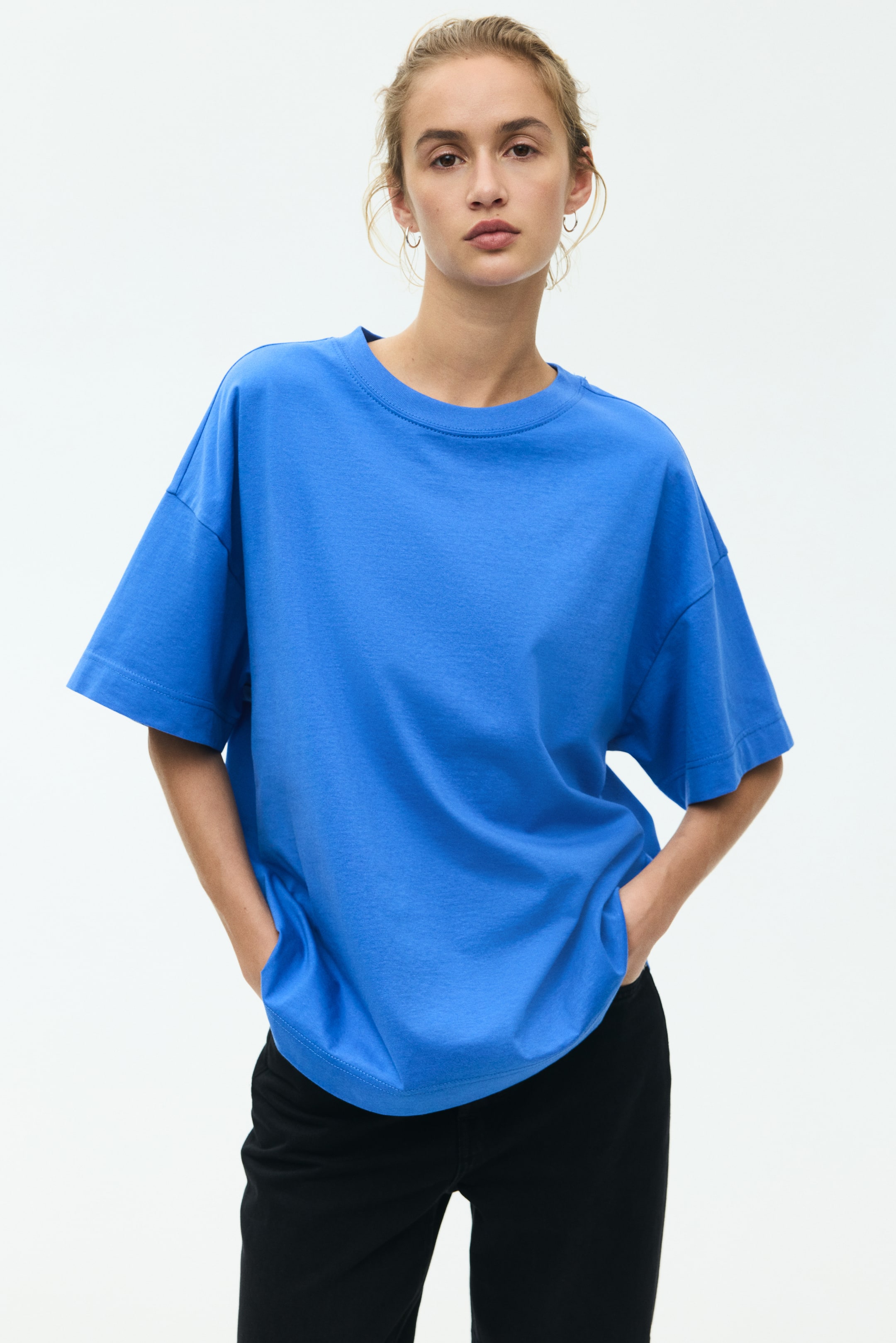 View larger image: The model is donning an oversized blue cotton jersey t-shirt, showcasing a rib-trimmed neckline, dropped shoulders, and a straight-cut hem, while placing her hands in the pockets of her black trousers.