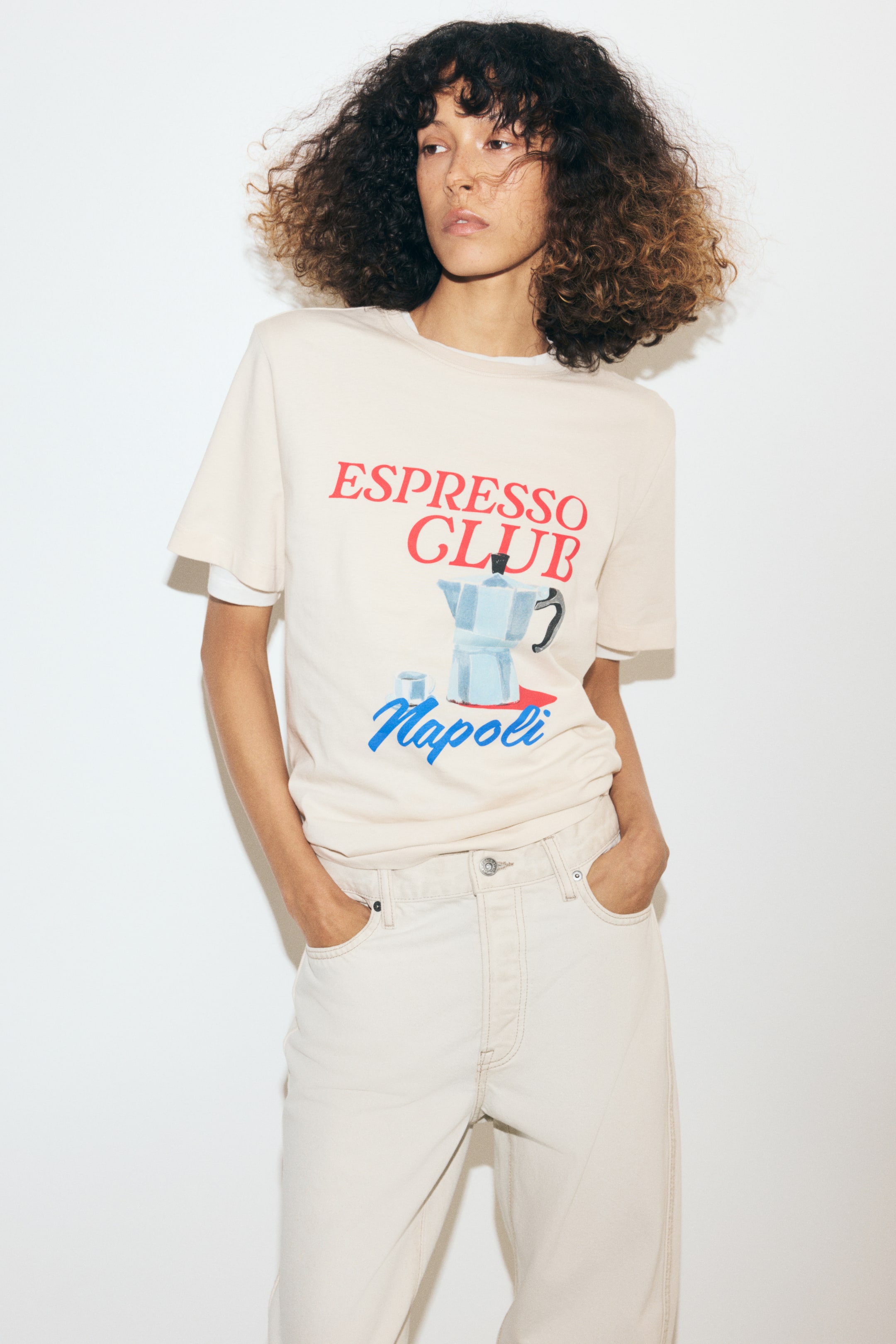 Ampliar la imagen: La modelo lleva puesta una camiseta beige claro con cuello redondo, hombros ligeramente caídos y un estampado frontal de una moka con el texto "ESPRESSO CLUB Napoli". La camiseta se lleva sobre una camiseta blanca y se combina con jeans de color claro.