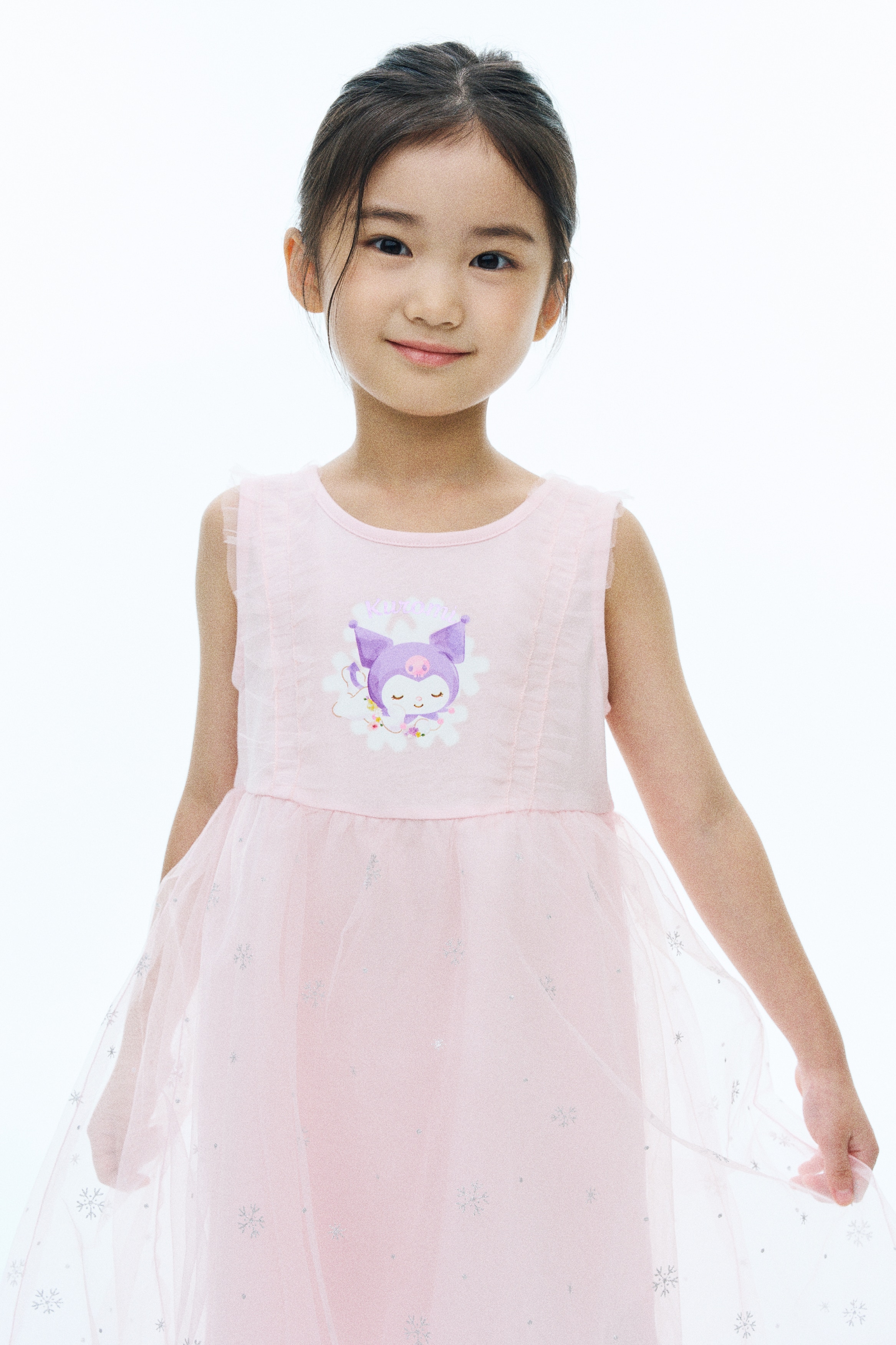 A light pink sleeveless dress is shown from the front, featuring a round neckline and a graphic of the character Kuromi sleeping on a cloud on the chest. The dress showcases sheer vertical tulle ruffles along the bodice and a layered tulle skirt decorated with silver glittery snowflakes.