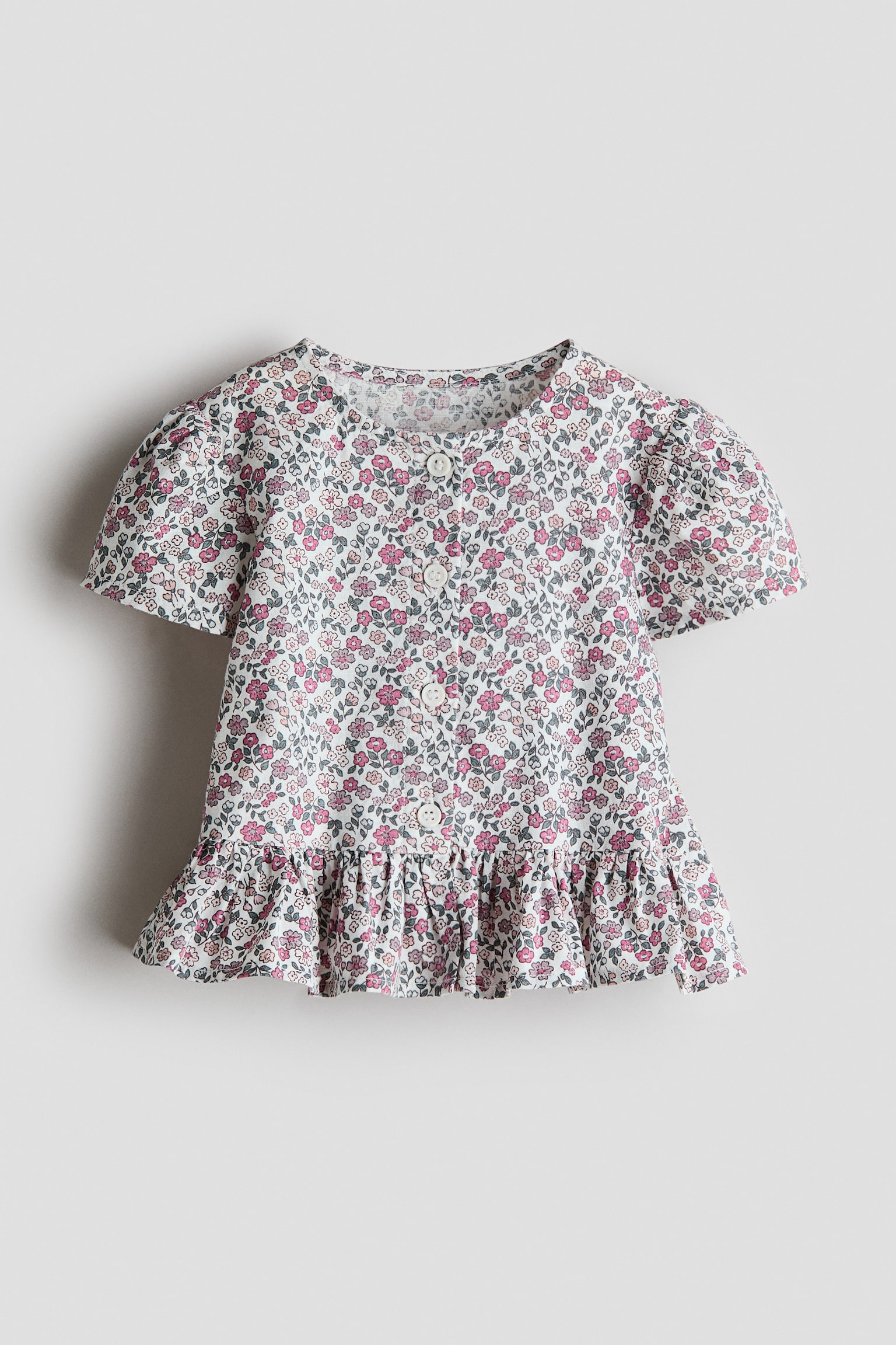 Blouse featuring an all-over floral print with pink, grey, and white flowers on a white background. It showcases a round neckline, a front button placket with four white buttons, short puff sleeves, and a flared, ruffled hem.