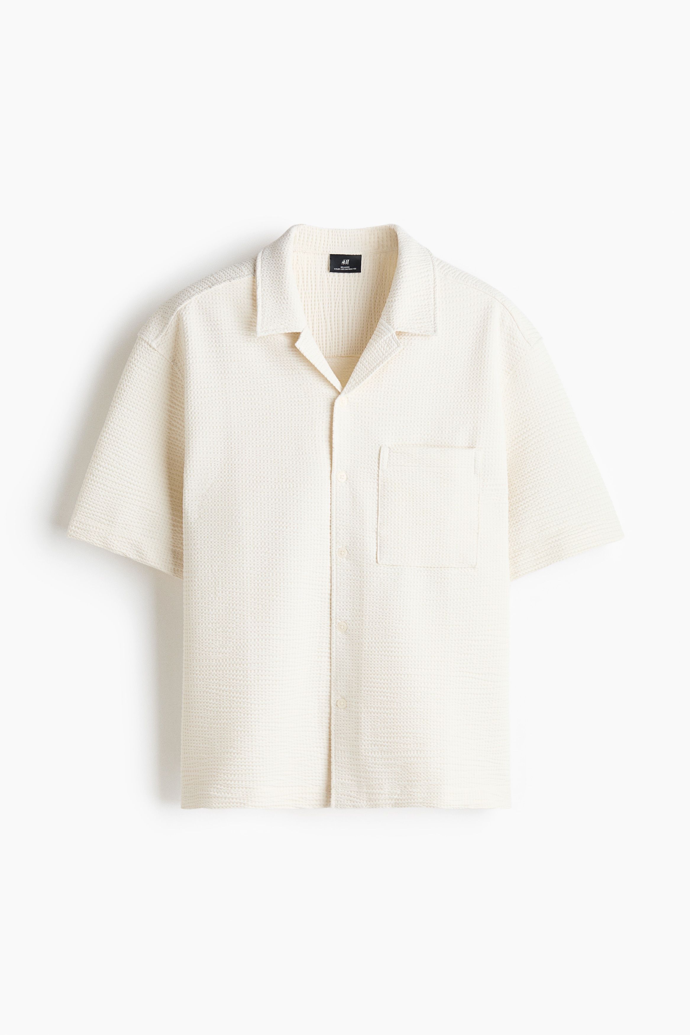 Une chemise Ã  manches courtes en mÃ©lange de coton Ã  texture gaufrÃ©e blanc cassÃ© est portÃ©e par le mannequin. La chemise prÃ©sente une coupe dÃ©contractÃ©e, un col camp, une seule poche poitrine et un ourlet droit, et elle est agencÃ©e avec un pantalon foncÃ©.