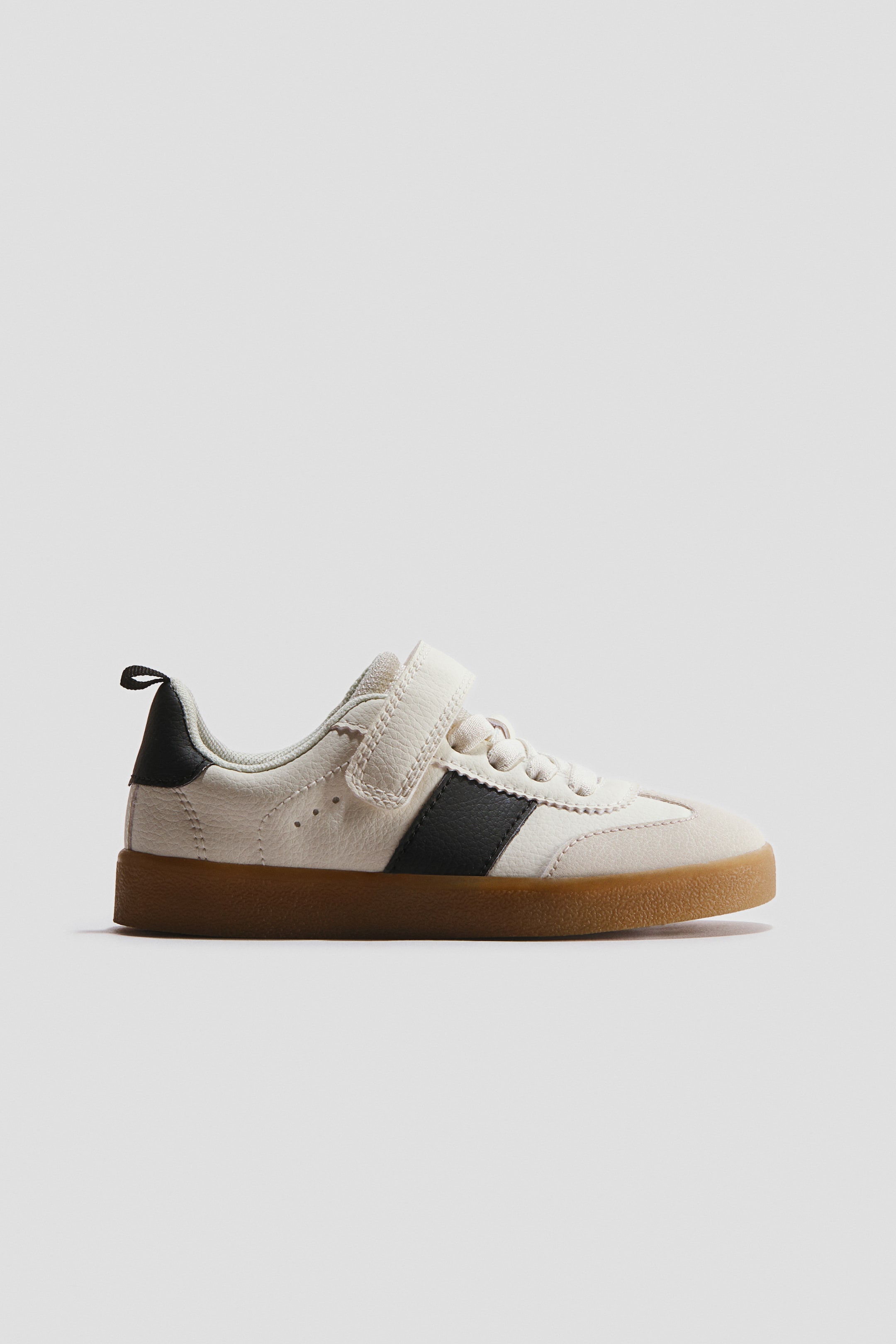 View larger image: The cream low-top sneaker showcases its side profile, boasting a textured upper adorned with two black stripes on the side panel and a brown gum rubber sole. A cream velcro strap secures the shoe over white laces, which are detailed with a serrated edge along the eyelets, and a black heel tab is visible.
