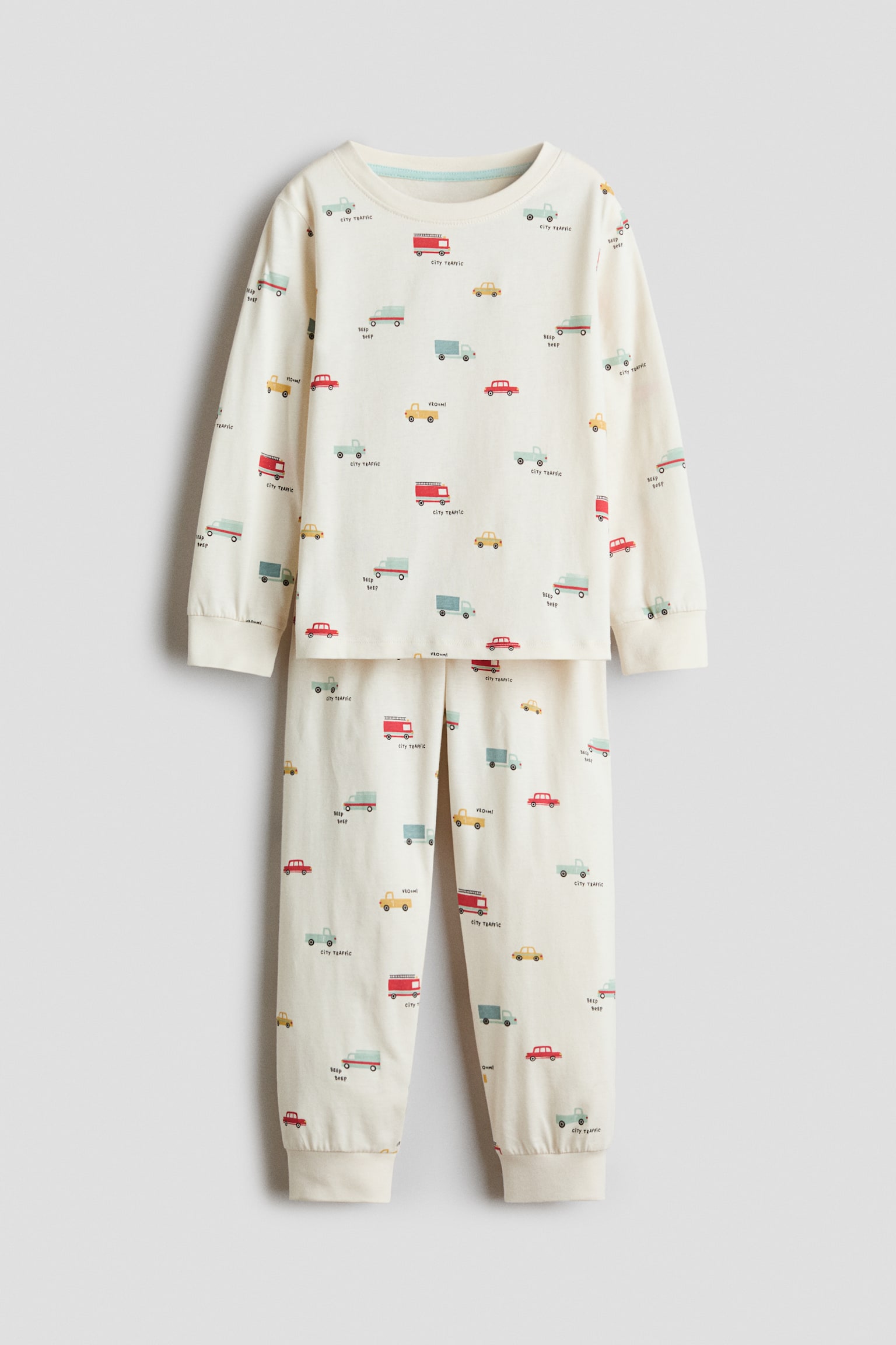 Un pigiama da bambino in jersey di cotone bianco sporco presenta una stampa all-over di veicoli giocattolo colorati. Mostrato frontalmente, il top a maniche lunghe presenta un girocollo rifinito a costine e polsini a costine, mentre i pantaloni coordinati mostrano orli alla caviglia a costine.