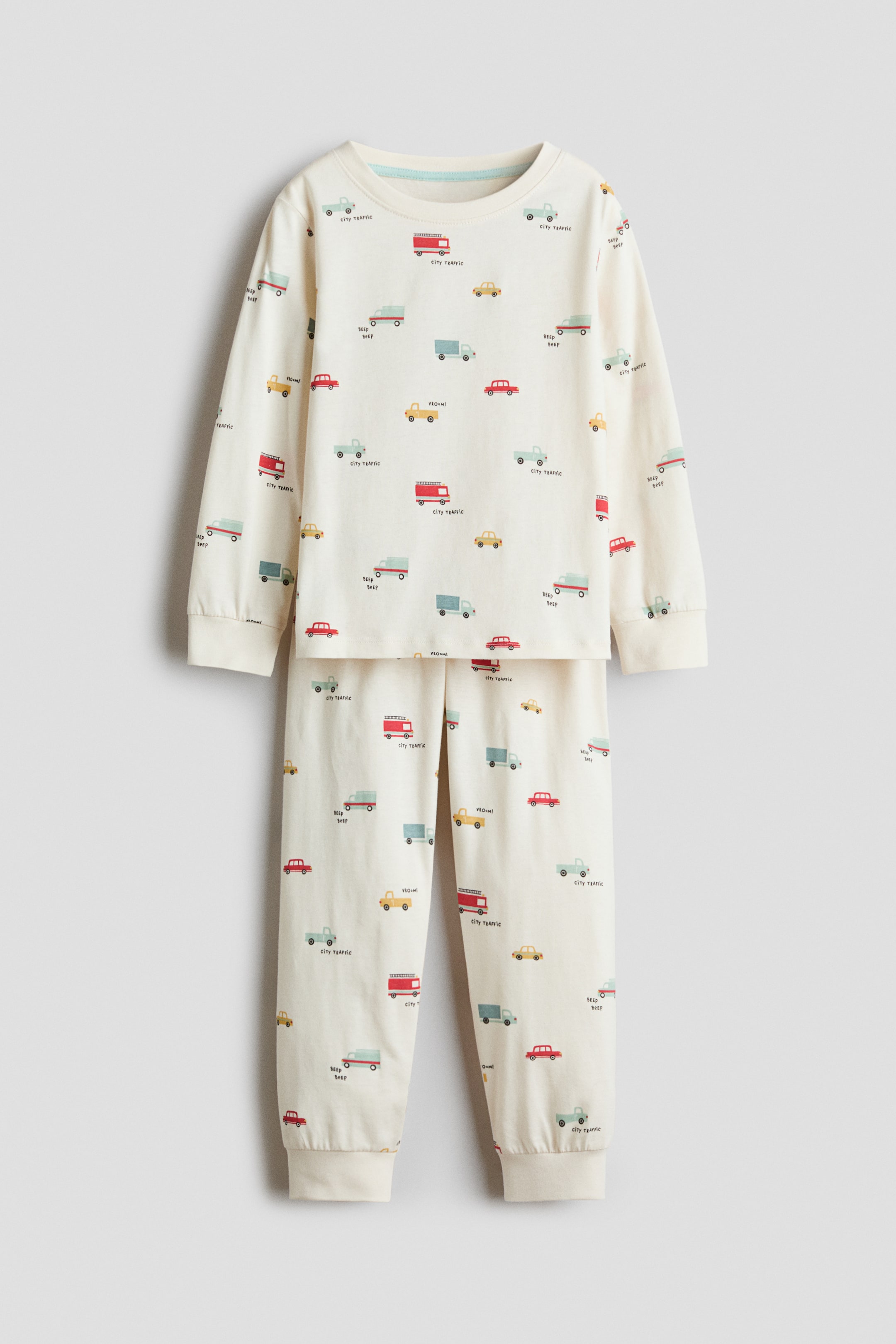 View larger image: A children’s pyjama set in off-white cotton jersey featuring an all-over print of colourful toy vehicles. Shown from the front, the long-sleeve top has a round, rib-trimmed neckline and ribbed cuffs, while the matching trousers have ribbed ankle hems.