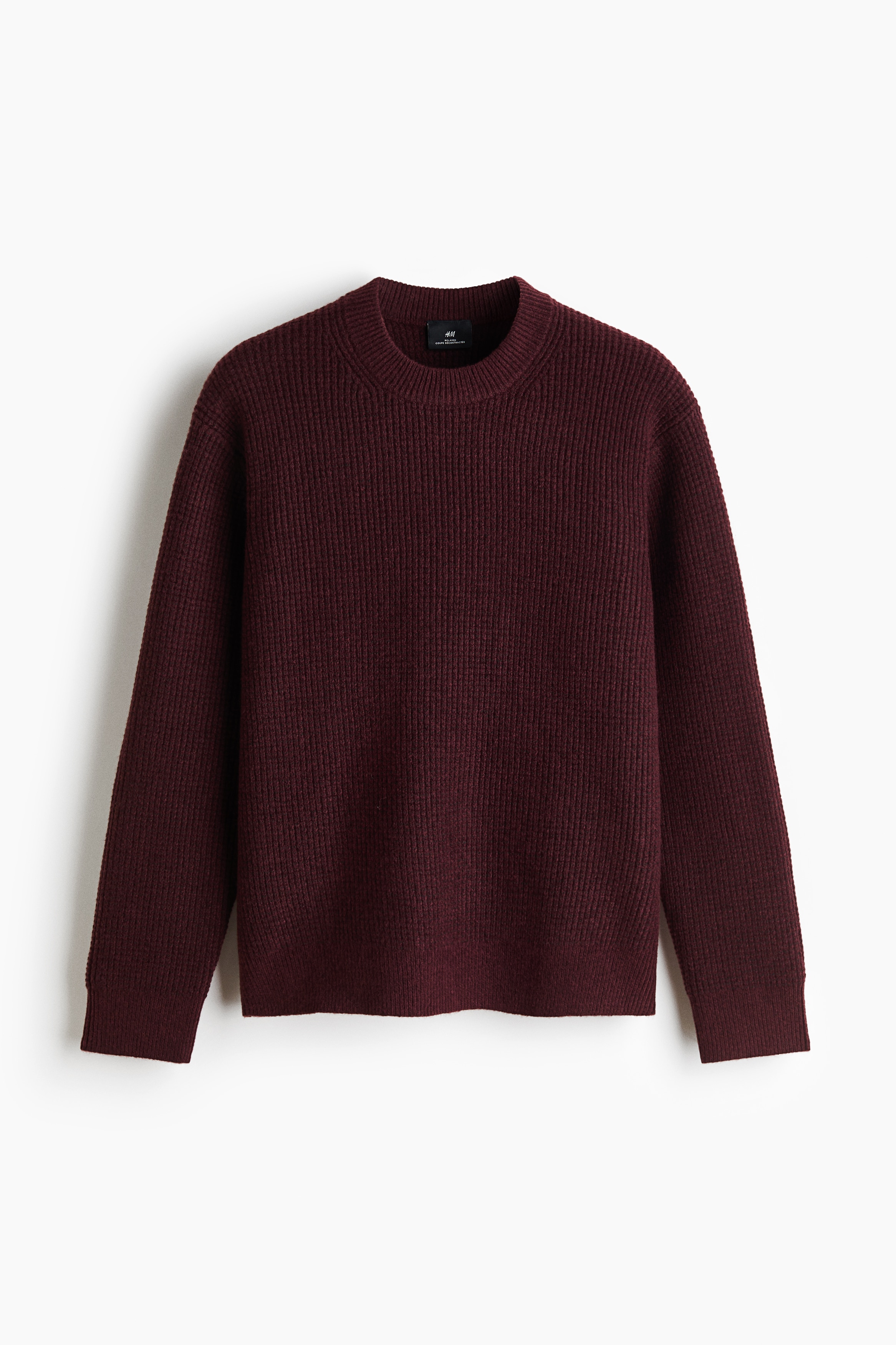 The model is wearing a burgundy textured knit sweater featuring a round ribbed neckline, ribbed cuffs, and a ribbed hem. The relaxed-fit sweater is layered over a white collar and paired with off-white trousers.