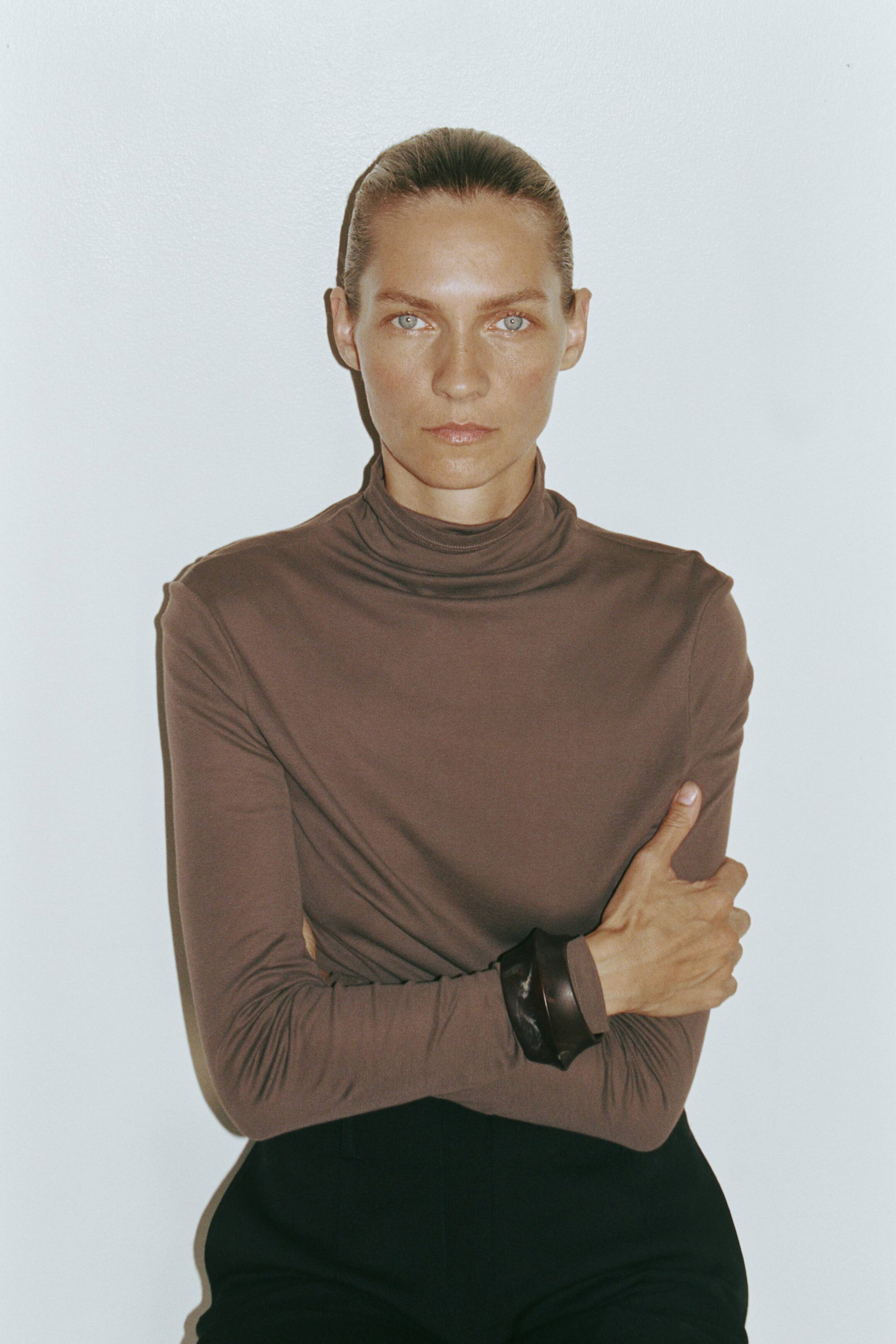 View larger image: A brown, high-neck jersey top with long sleeves is being worn by the model, who is posing with crossed arms. The top is tucked into black trousers and styled with a wide, dark cuff bracelet over one sleeve.