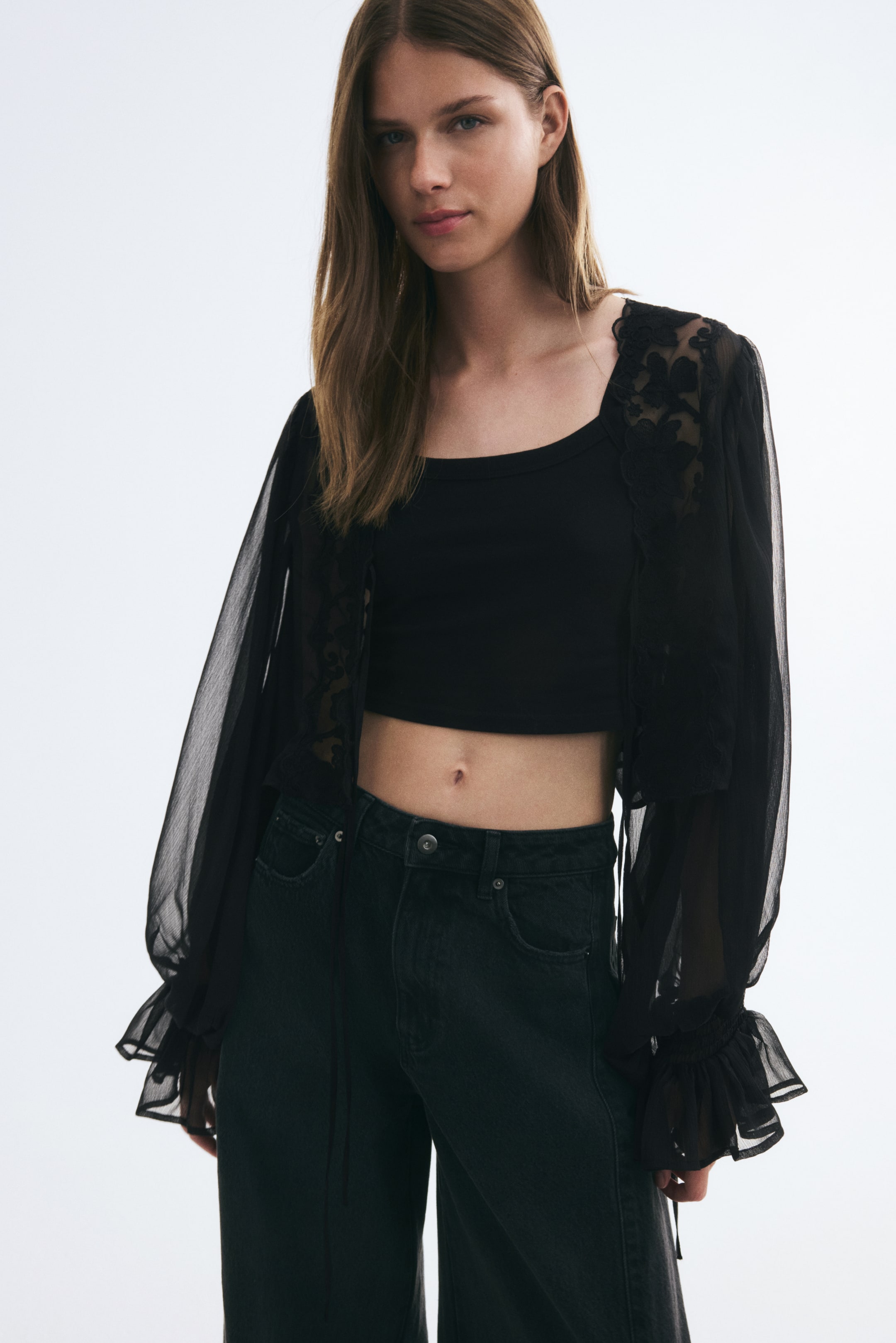 View larger image: A black crinkled chiffon blouse with floral lace trim is being worn open by the model, layered over a black crop top and dark jeans. The sheer blouse features long balloon sleeves that end in elasticized, flared ruffled cuffs.