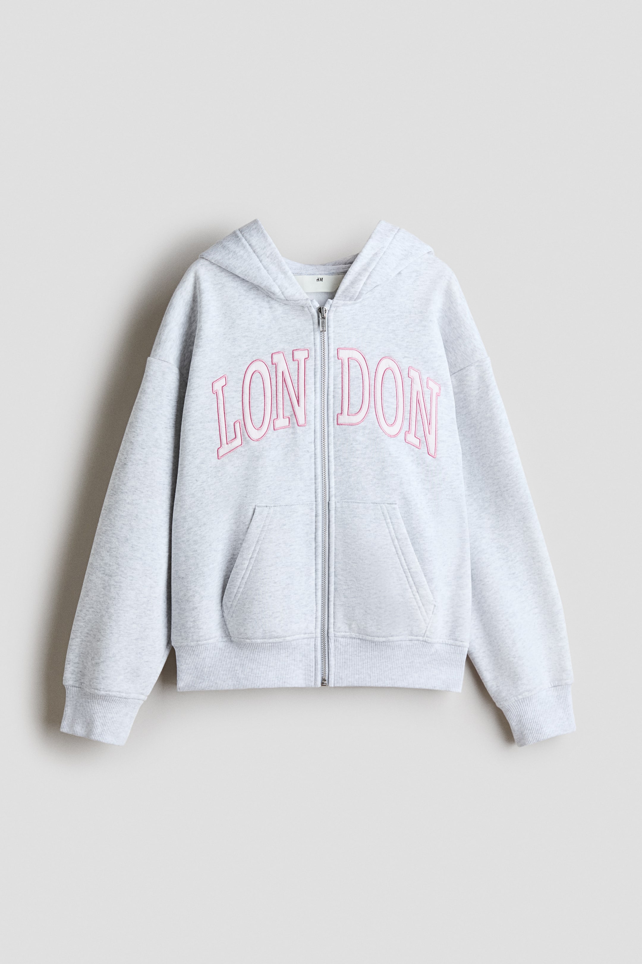 View larger image: A light gray marl hoodie is shown from the front, featuring a full-length zipper, a hood, and a kangaroo pocket. The word 'LONDON' is embroidered across the chest in pink with a white outline, and it has ribbed cuffs and a ribbed hem.