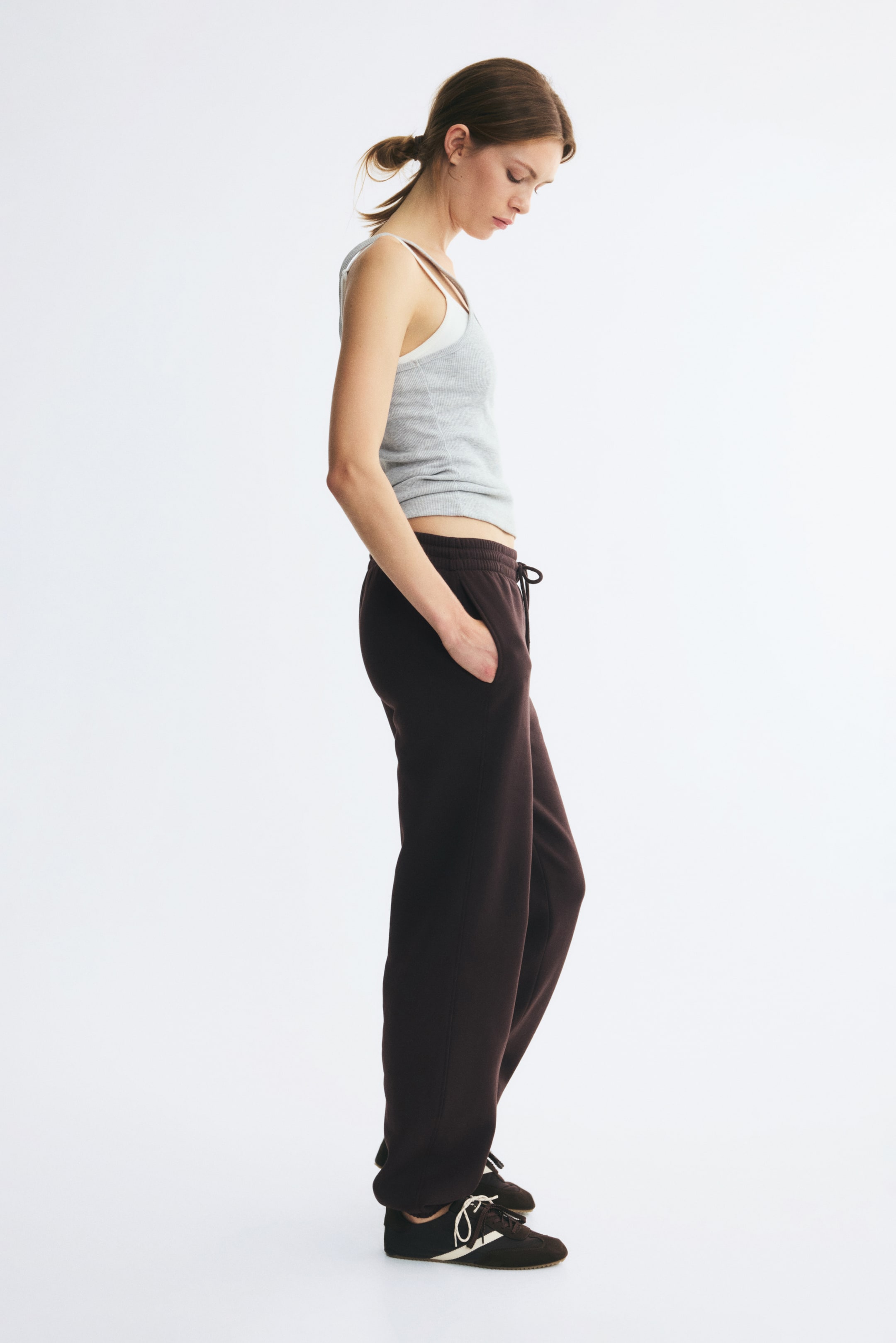 View larger image: Track pants in a dark brown sweatshirt fabric are worn by the model, who is standing in profile with one hand resting in the side pocket. The high-waisted pants feature an elasticated waistband with a drawstring and gathered elastication at the hems, styled with a grey tank top and dark sneakers.