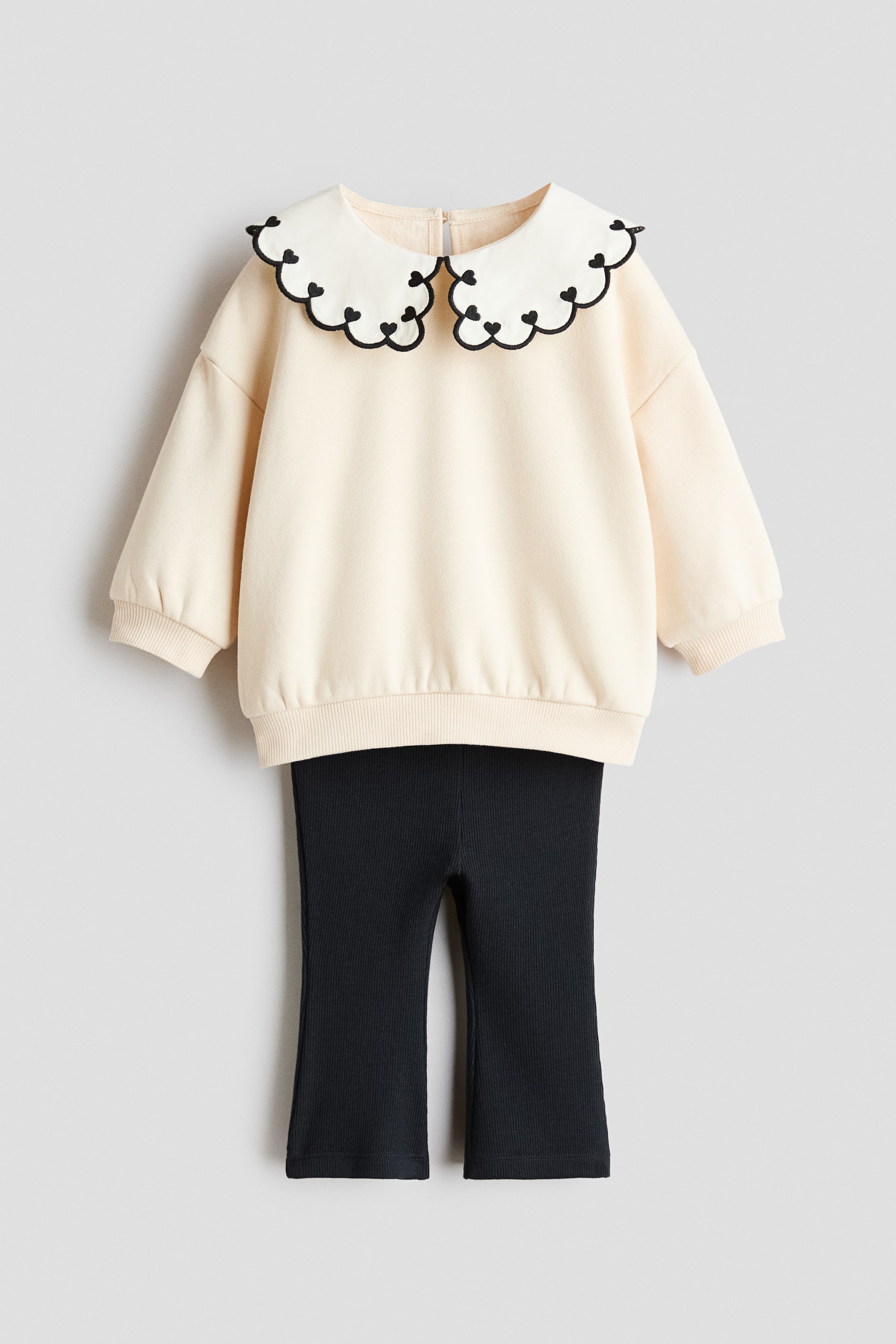 Se større billede: Et sæt, FRONT, præsenterer en cremefarvet top i blødt sweatshirtstof med en stor dobbeltkrave med bølgede kanter og broderede sorte hjerter, sænkede skuldre, ribbede manchetter og en ribbet kant, kombineret med sorte leggings i blød, ribbet jersey med vidde ben.