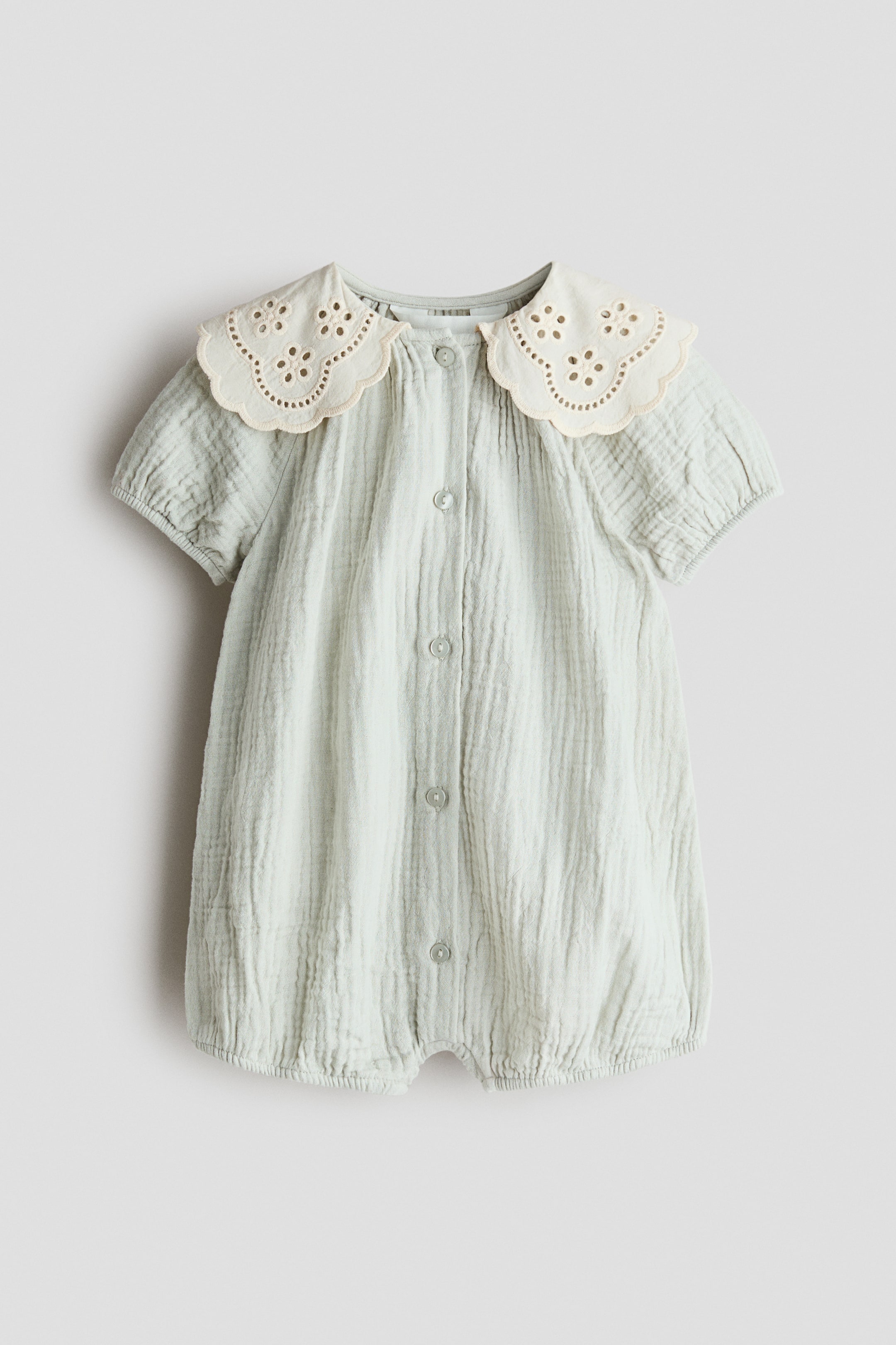 View larger image: A light green muslin romper suit is showcased from the front, highlighting a round neckline and a cream-coloured, scalloped collar adorned with broderie anglaise. It is crafted with a full button-down front, short puffed sleeves with elasticated cuffs, and elasticated leg hems.