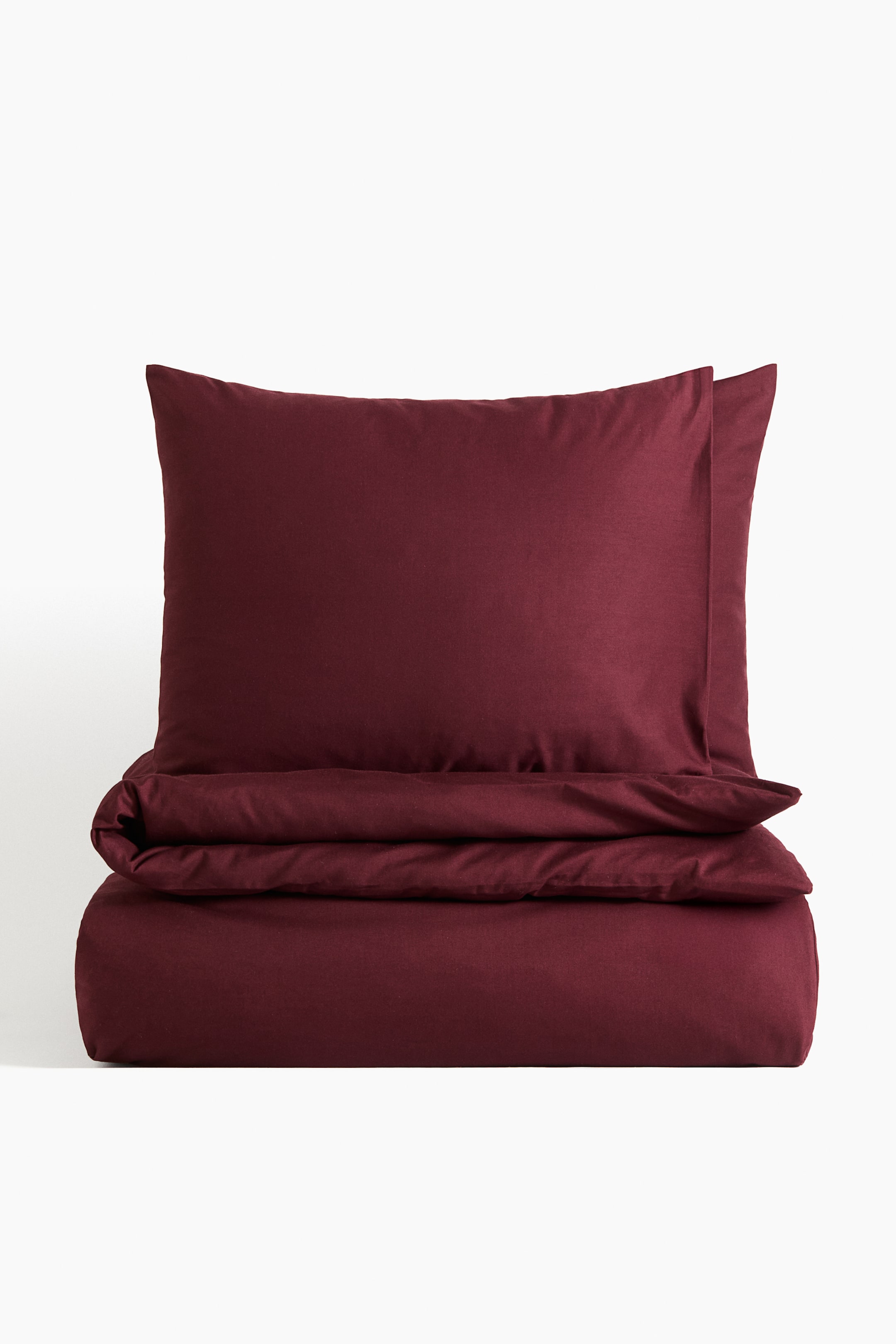 View larger image: A deep red cotton duvet cover set is being presented. The duvet cover is lying folded horizontally at the front, and two matching pillowcases are resting stacked on top.