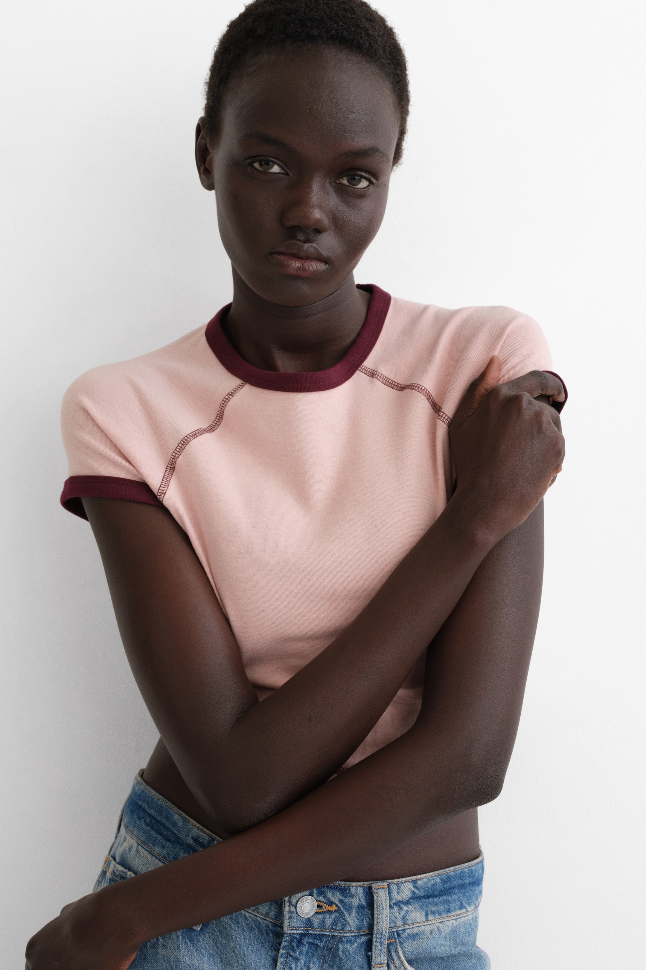 View larger image: A light pink ringer T-shirt featuring a crew neck, short raglan sleeves and a cropped fit, finished with dark red trim at the collar and sleeve cuffs, plus visible contrast stitching along the raglan seams.