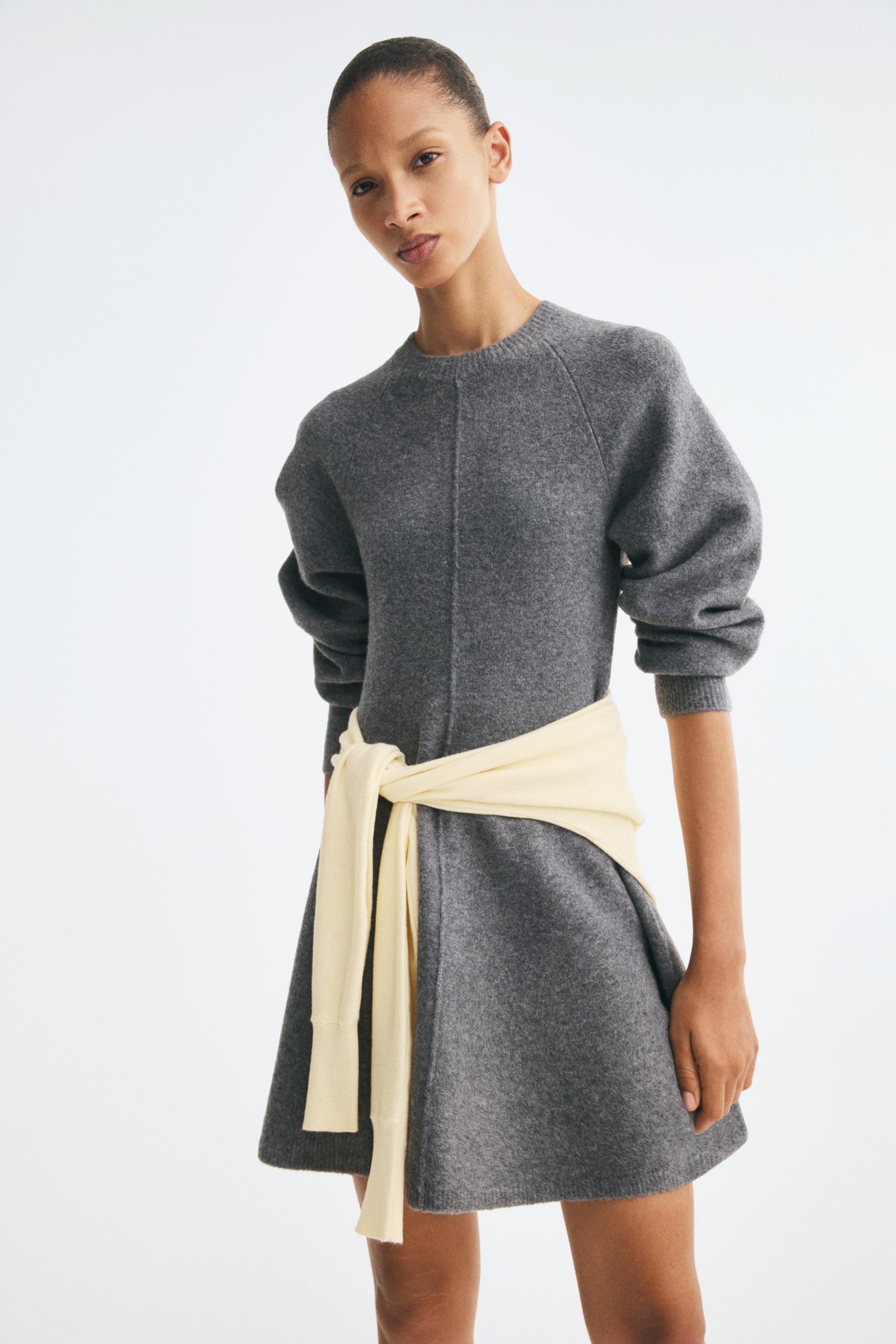 View larger image: A short grey knitted dress is shown from the front, featuring a round rib-trimmed neckline, long raglan sleeves, a flared skirt, and a prominent vertical seam running down the centre. A light yellow jumper is tied around the waist, accentuating the dress's fitted silhouette.