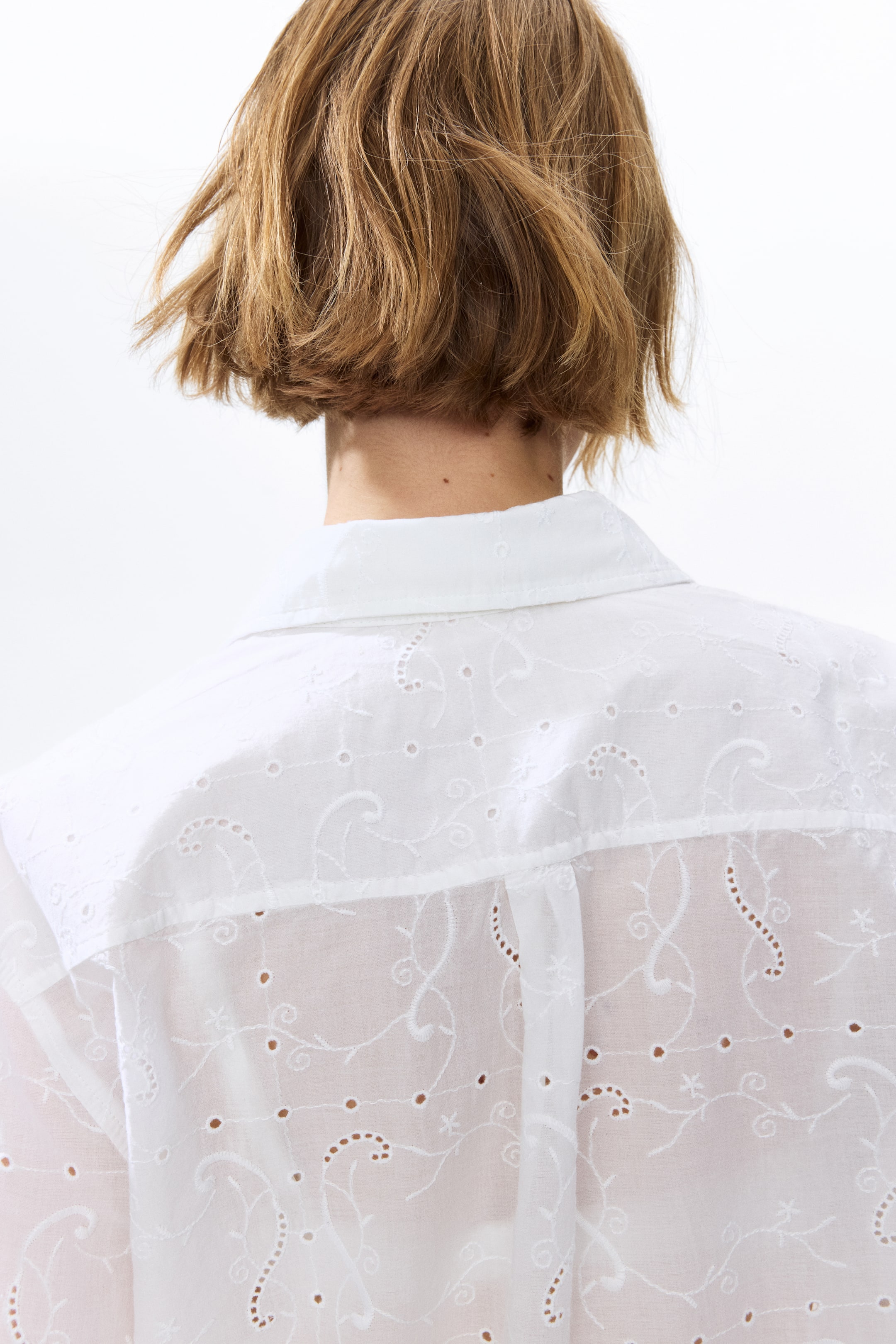 View larger image: The model is wearing an oversized white cotton shirt with broderie anglaise, viewed from the back. The shirt comes with a collar, a double-layered yoke, and a box pleat at the center back.