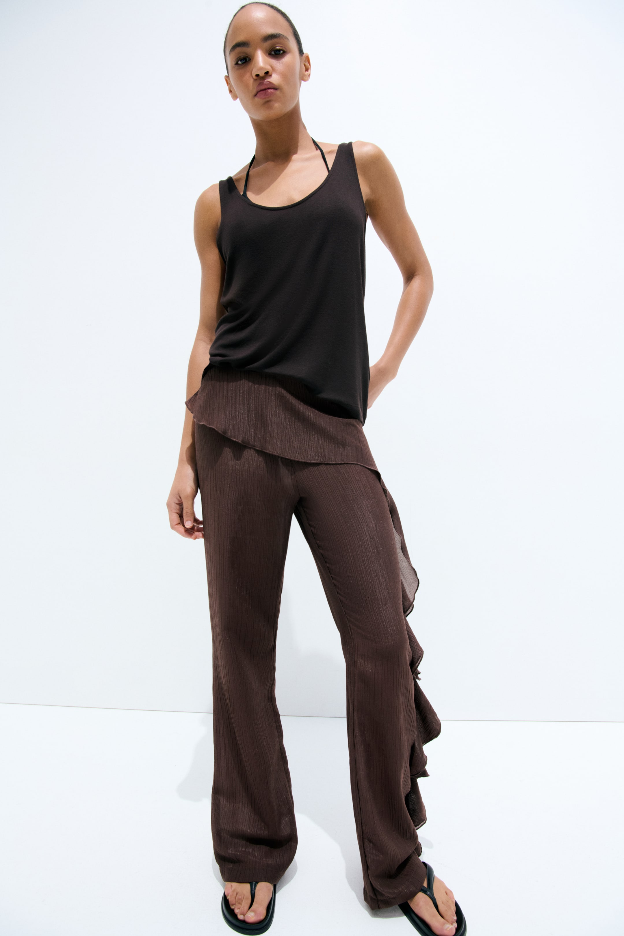Voir une image plus grande: Pantalon marron à jambes larges avec une texture verticale fine et des détails asymétriques en volants le long de la jambe droite et de la taille, tandis qu'un débardeur à encolure dégagée foncée est superposé à une bretelle de licou noire visible.