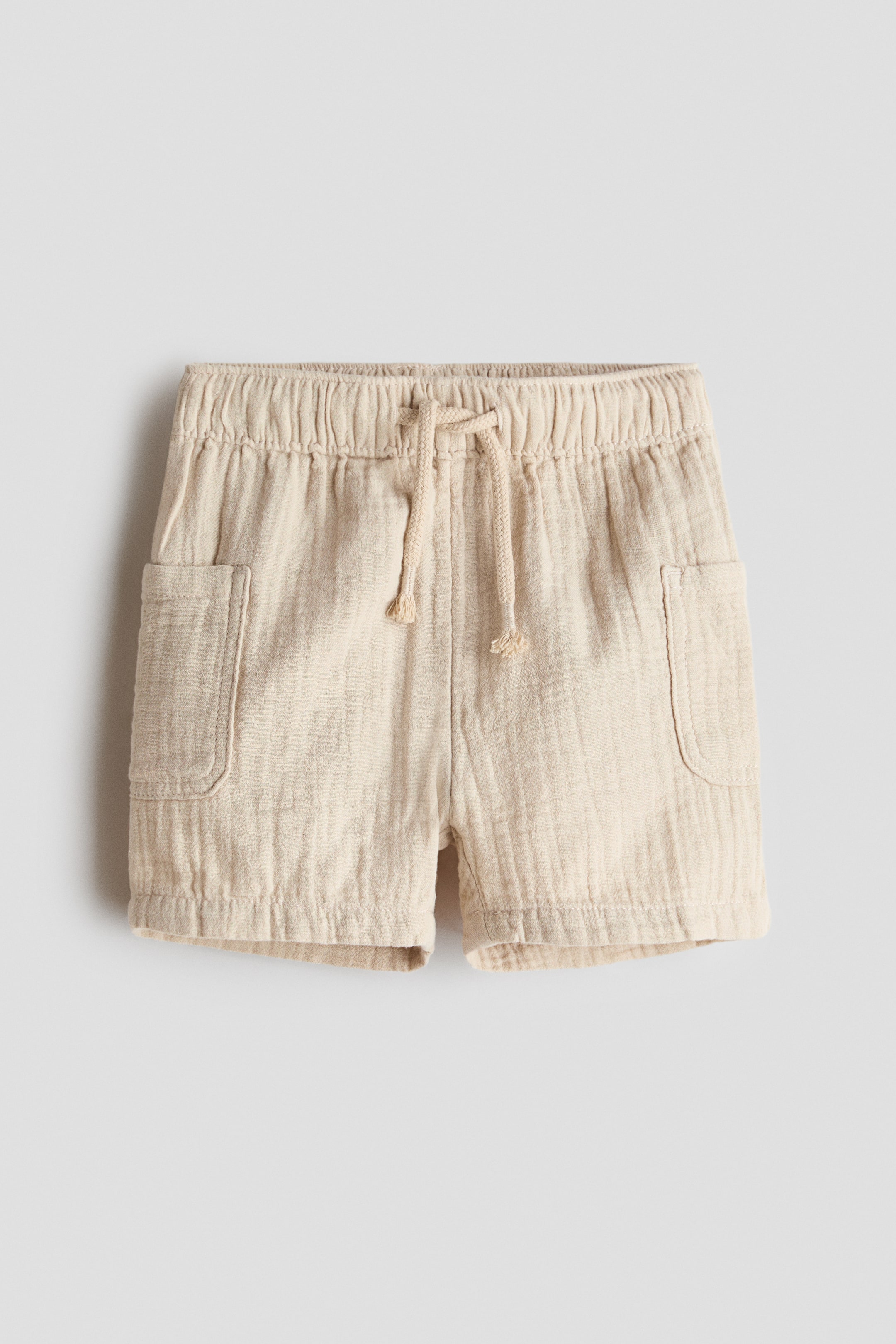 View larger image: The light beige shorts feature an elastic waistband with a drawstring tied at the front, and two front patch pockets with rounded corners are visible on each side. The shorts are crafted from a crinkled fabric.