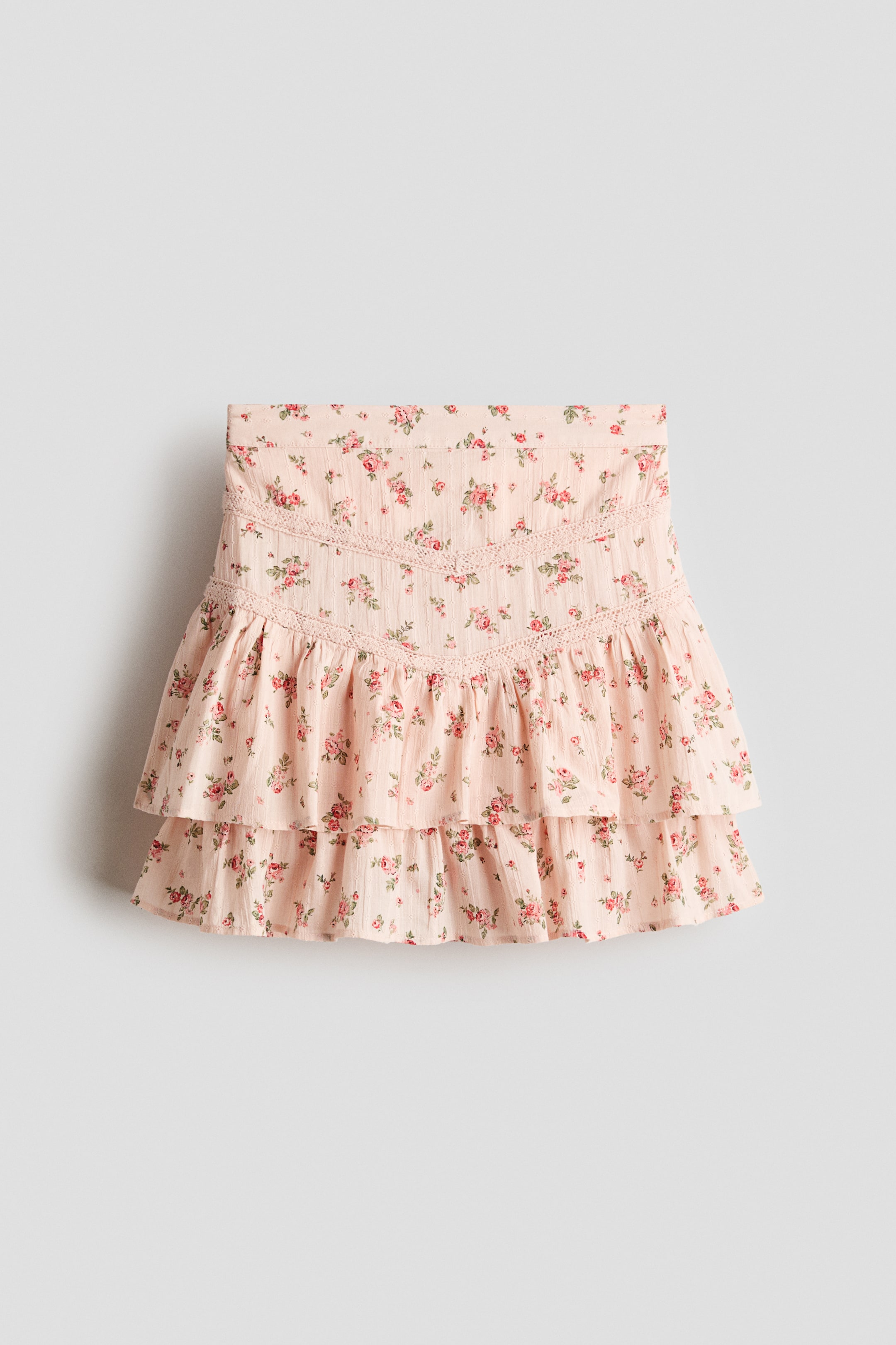 Grotere afbeelding bekijken: Een uitlopende rok toont zijn voorkant, gemaakt van een luchtige katoenen weefsel met een lichtroze bloemenprint, voorzien van V-vormige kanten inzetstukken en twee geplooide lagen.
