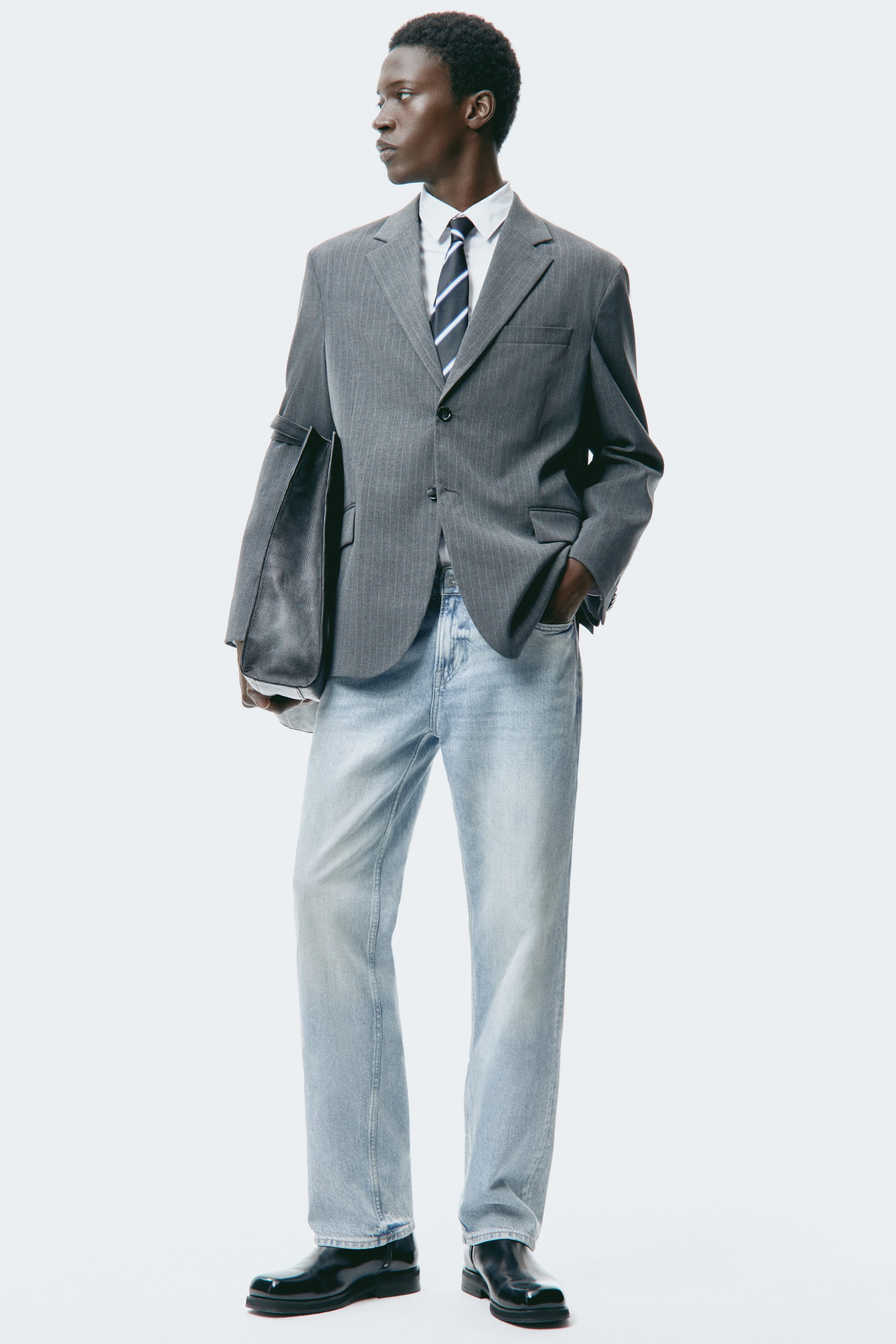 Voir une image plus grande: Une personne porte un blazer gris à fines rayures, coupe décontractée, à revers crantés et fermeture à deux boutons, superposé à une chemise blanche à col et à une cravate rayée. Elle porte également un jean denim à lavage clair et des chaussures en cuir noir poli, et tient un sac fourre-tout noir sur son bras gauche.