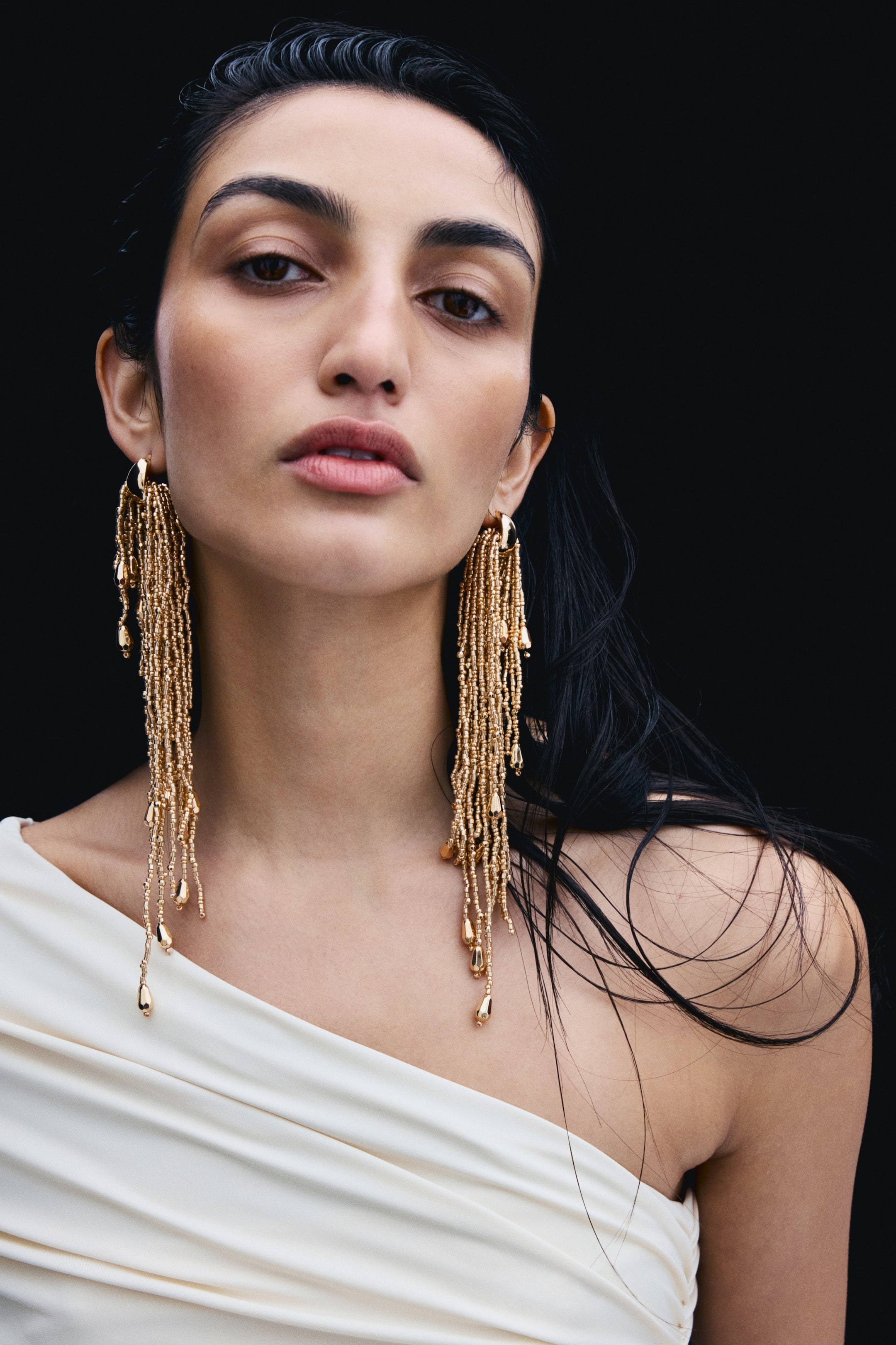 View larger image: Gold-toned dangling earrings with a small metal hoop top and long, cascading strands of tiny beads are being worn by the model. The shoulder-length strands, ending in small teardrop-shaped beads, drape towards the collarbone against a white off-the-shoulder top.