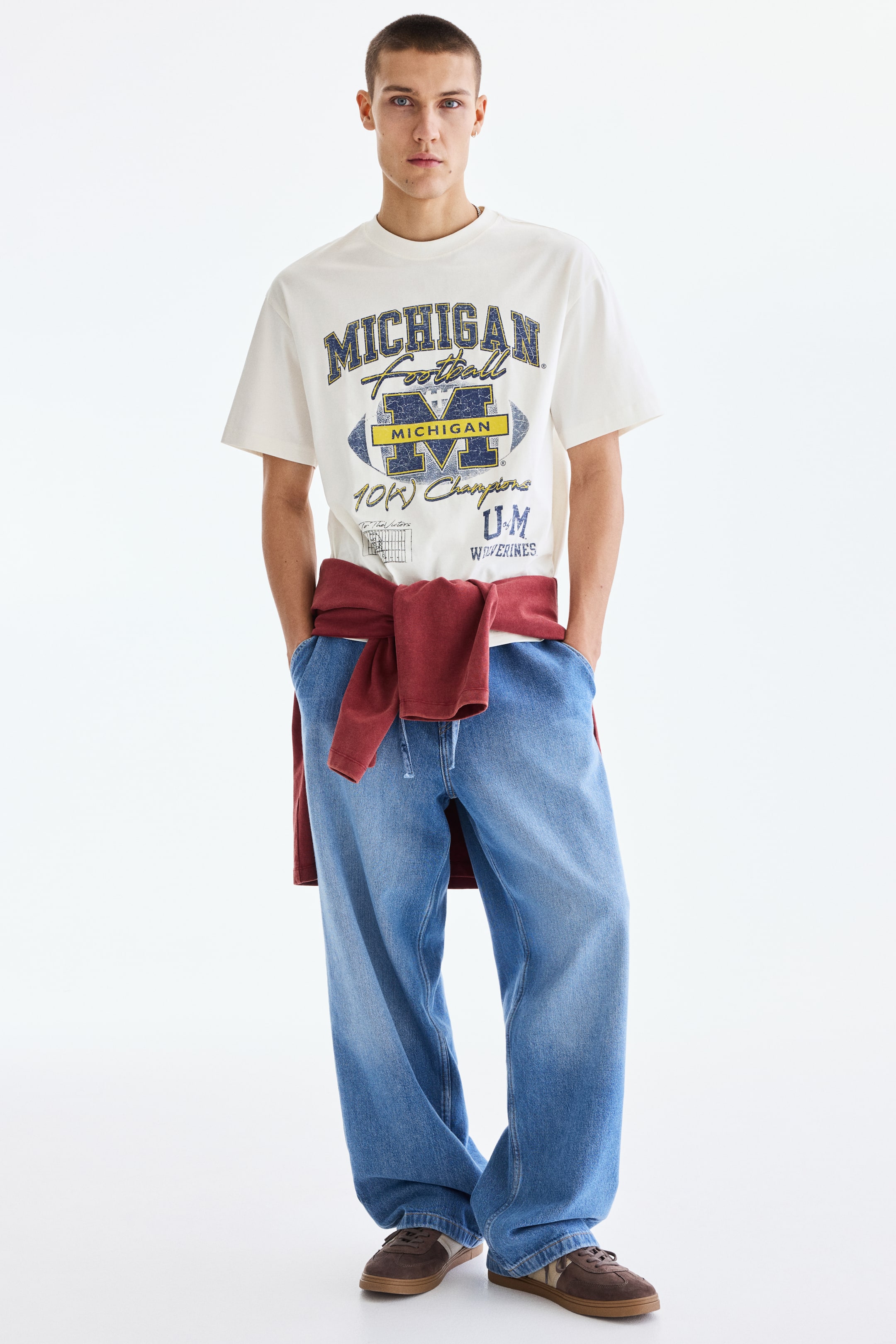 View larger image: A short-sleeved t-shirt in off-white showcases a distressed "MICHIGAN Football" graphic in blue and yellow, prominently displayed on the chest. The design highlights a large "M" within a football shape, accompanied by the text "10K Champions," "U.M.," and "Wolverines" below.