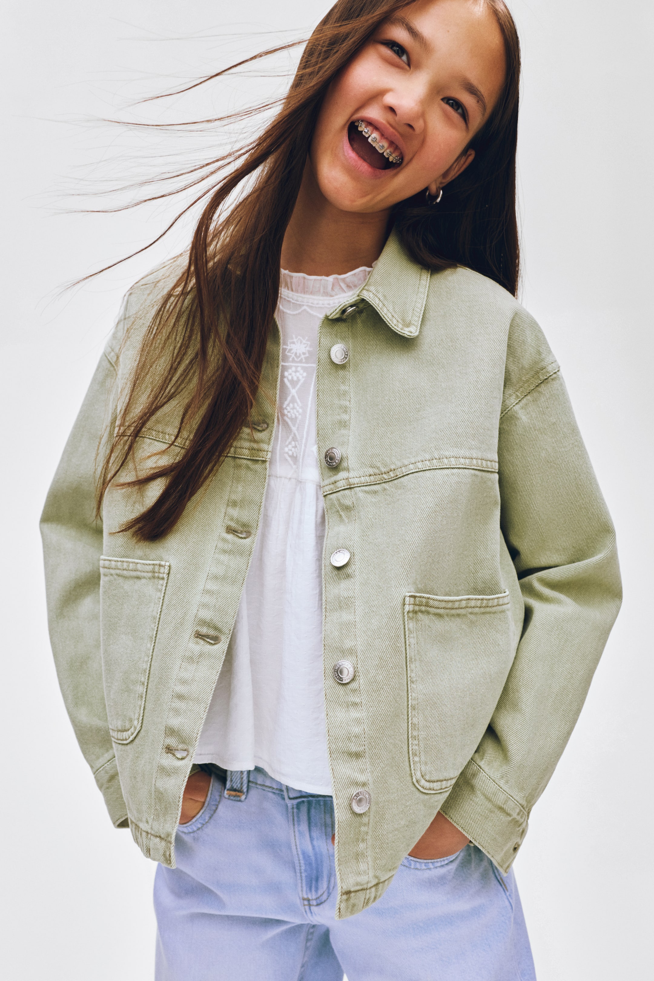 Ver imagen más grande: Una chaqueta vaquera de color verde claro se lleva abierta sobre una blusa blanca y unos vaqueros azul claro. Presenta un cuello en punta, botones plateados en la parte delantera, dos bolsillos de parche en el pecho con solapas y dos bolsillos de parche más grandes en la parte inferior.