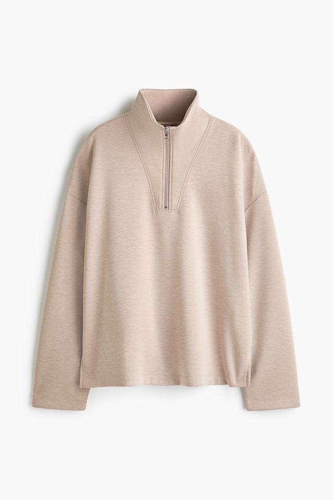 En lys beige half-zip-genser vises forfra, med hÃ¸y oppstÃ¥ende krage med sÃ¸lvfarget metallglidelÃ¥s og dekorative V-formede sÃ¸mmer pÃ¥ brystet. Plagget har en avslappet passform med senkede skuldre, lange rette ermer og en pent avsluttet kant med smÃ¥ splitt i sidene. - 1