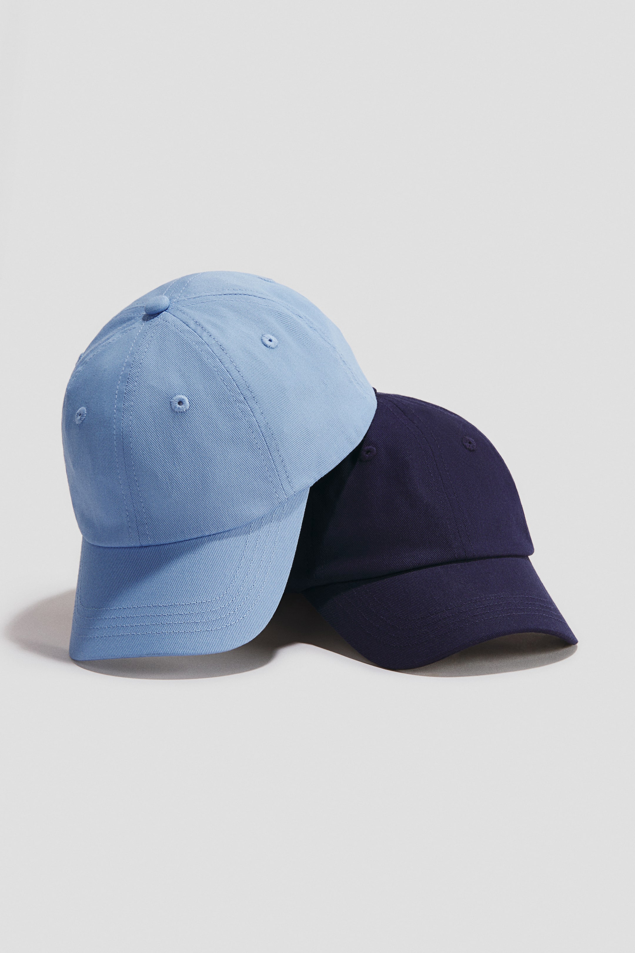 View larger image: Presenting two cotton twill caps: a light blue cap positioned in front of a navy blue cap. Both caps showcase a six-panel design, a top button, embroidered eyelets, and a curved brim with parallel stitching.