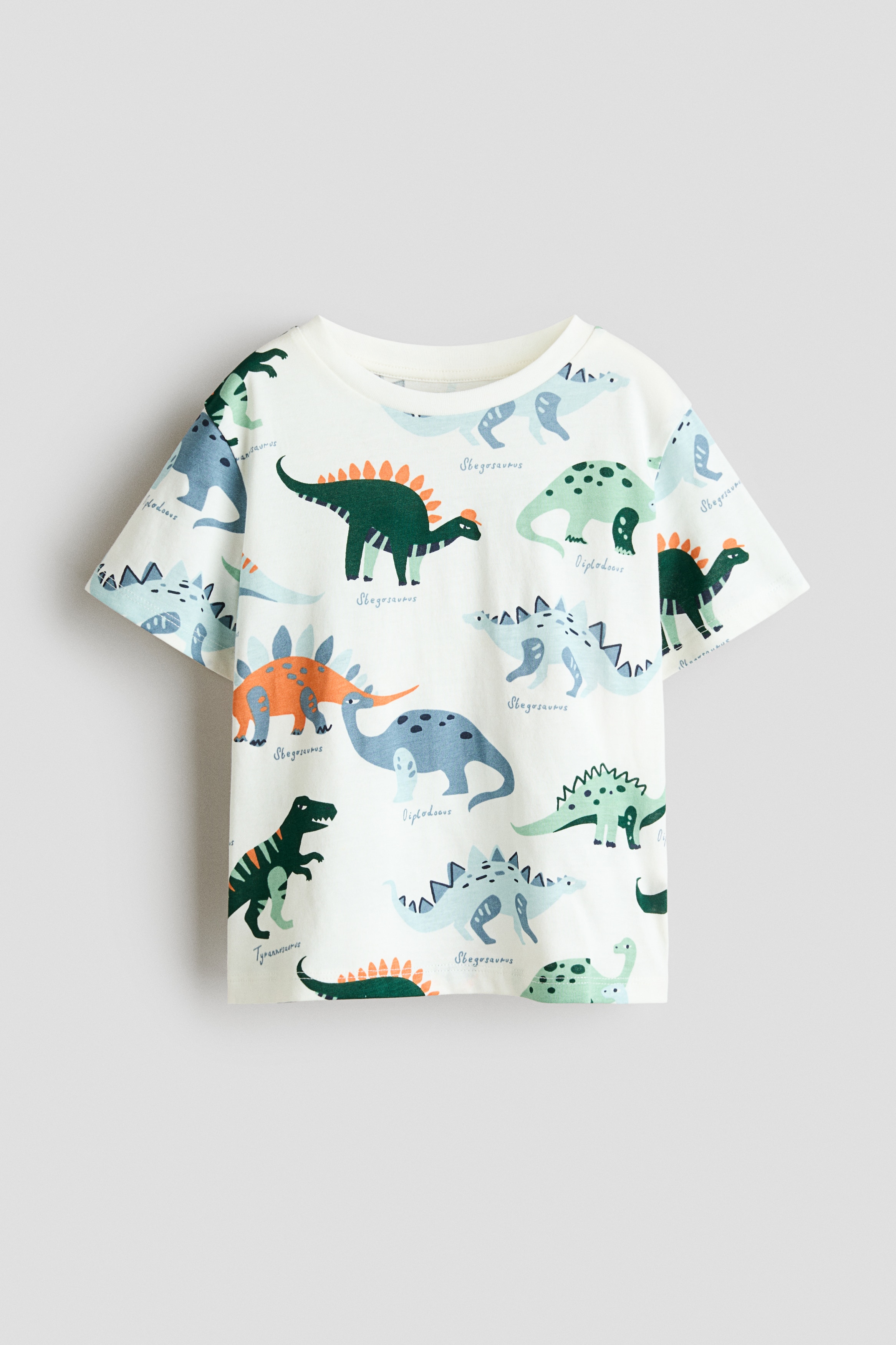 The model is wearing a white short-sleeved tee featuring a colourful all-over dinosaur print and a rib-trimmed round neckline. The tee is styled with loose, light beige cargo trousers and sneakers.