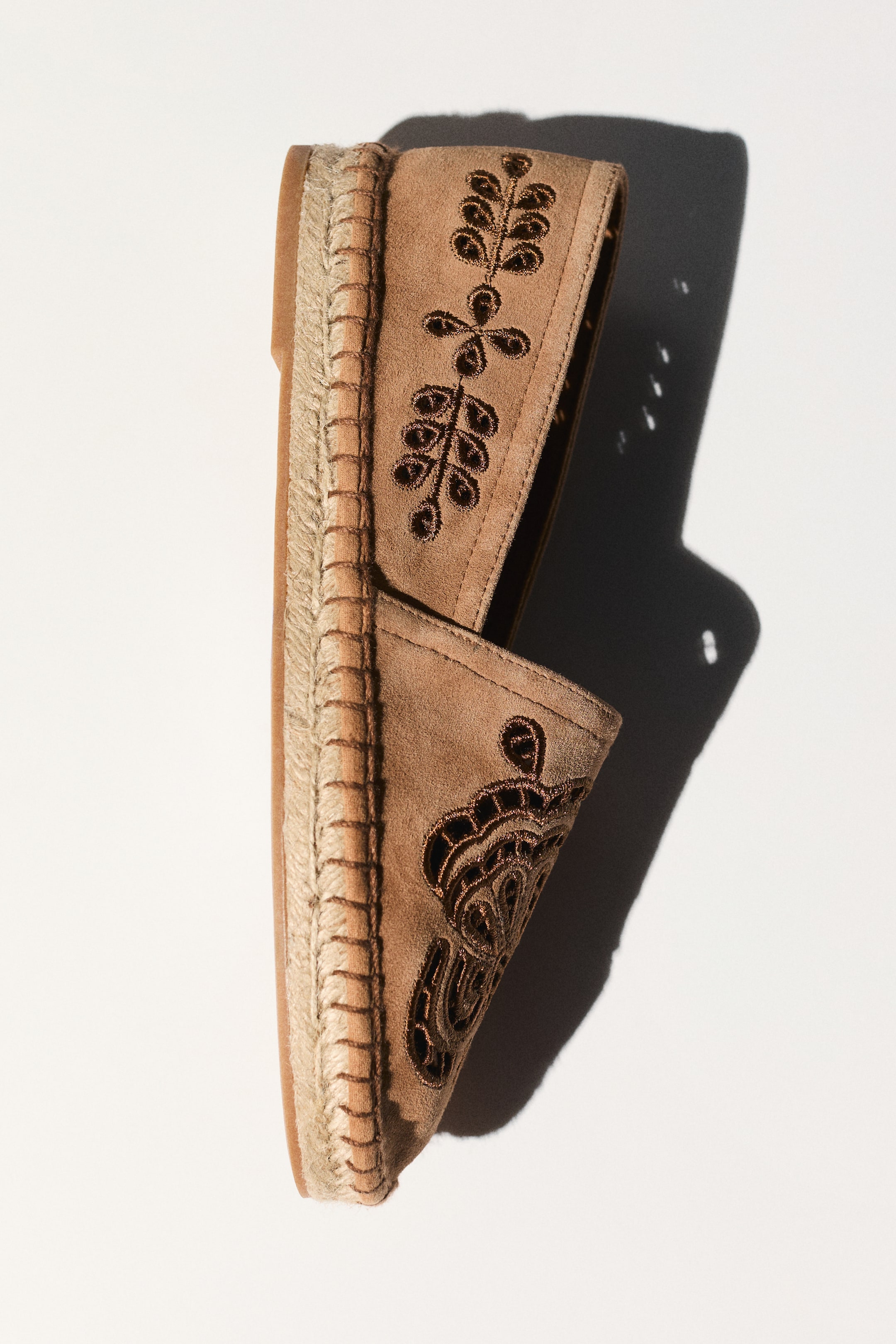 Visa större bild: En solbrun espadrillosko är vinklad diagonalt och visar upp sin flätade jutesula och en solbrun mockaöverdel. Överdelen har mörkbruna broderade blad- och blommönster.