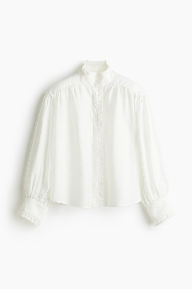 A white blouse is shown from the front, featuring a high ruffled stand-up collar, a button-front placket with ruffled trim, long voluminous sleeves, and wide ruffled cuffs. - 1