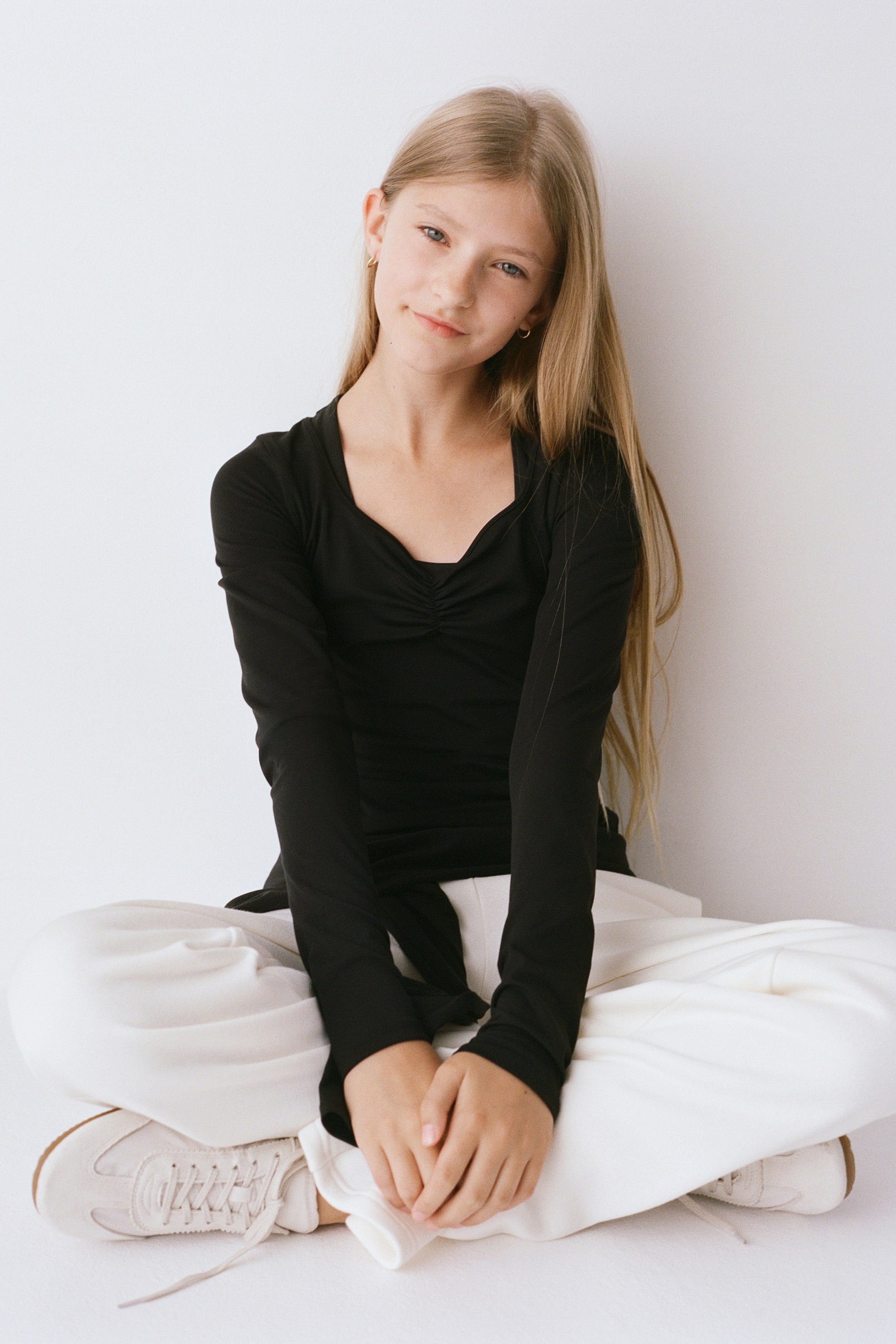 Grotere afbeelding bekijken: Een zwarte top met lange mouwen heeft een sweetheart-halslijn met gerimpelde details in het midden en een aansluitend silhouet, weergegeven vanuit een frontale hoek. De top wordt gestyled met losvallende witte broeken om de nauwsluitende pasvorm en lengte te benadrukken.