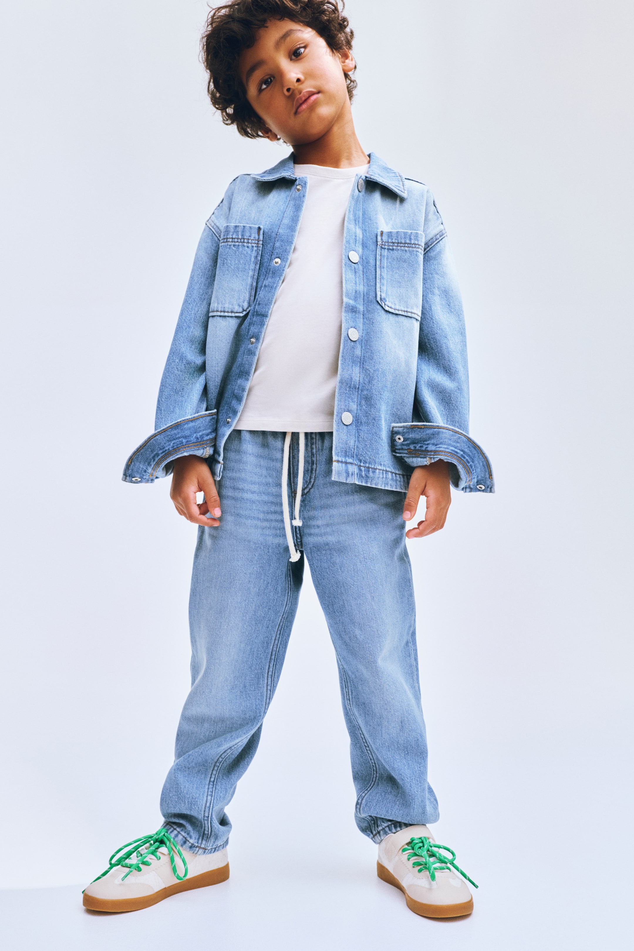 Zobrazit větší obrázek: Dětský outfit z lehké džínoviny je prezentován zepředu. Bunda je nošena otevřená přes bílé tričko, obsahuje límec, přední zapínání na knoflíky a dvě náprsní kapsy s klopami, zatímco rukávy jsou vyhrnuté; k tomu jsou nošeny odpovídající džínové kalhoty s bílou šňůrkou v elastickém pase a elastickými kotníky, doplněné světlými teniskami se zelenými tkaničkami.