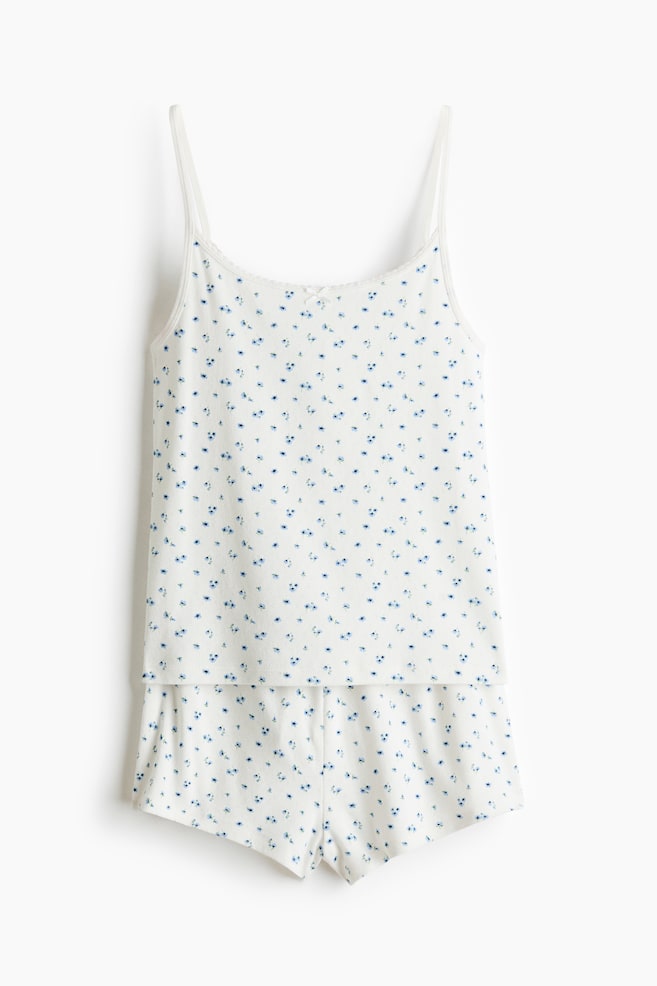 Een wit pointelle jersey pyjamaset wordt frontaal weergegeven, met een kleine blauwe bloemenprint. Het camisoletopje is ontworpen met smalle schouderbandjes, een ronde halslijn met een klein strikje, en wordt gecombineerd met bijpassende shorts met een elastische tailleband en een korte snit. - 1
