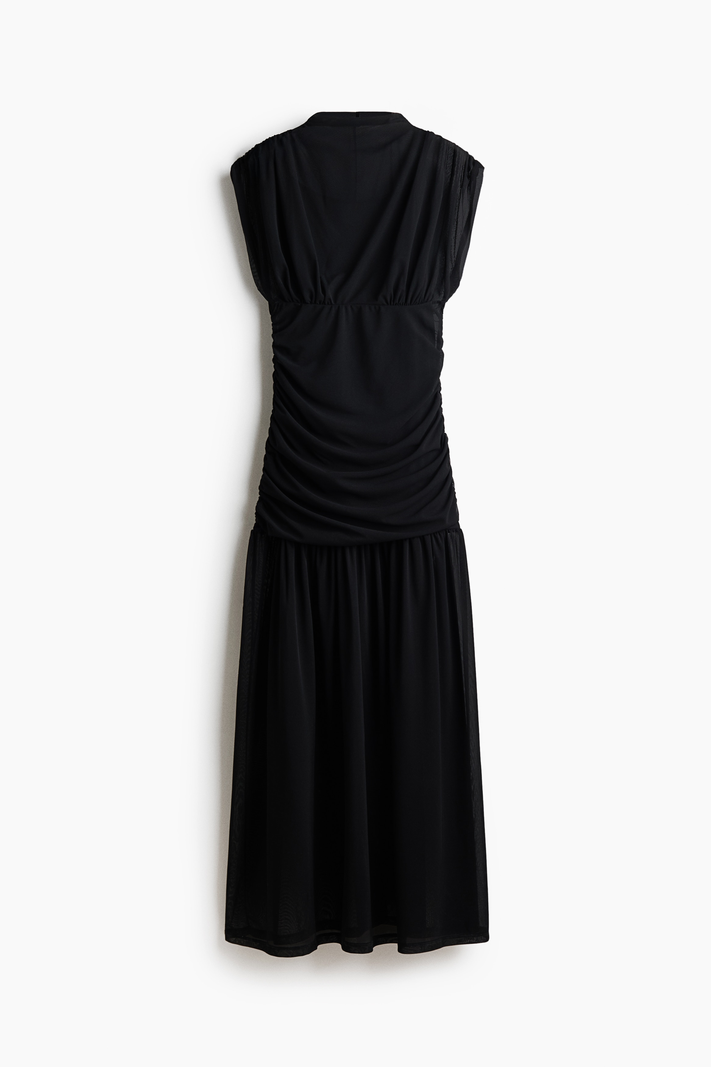 The model is wearing a black sleeveless maxi dress featuring a high neckline, a fitted bodice with horizontal ruching, and a sheer, draped maxi skirt. The sheer outer layer reveals a shorter underdress underneath, and the look is completed with black flat shoes.