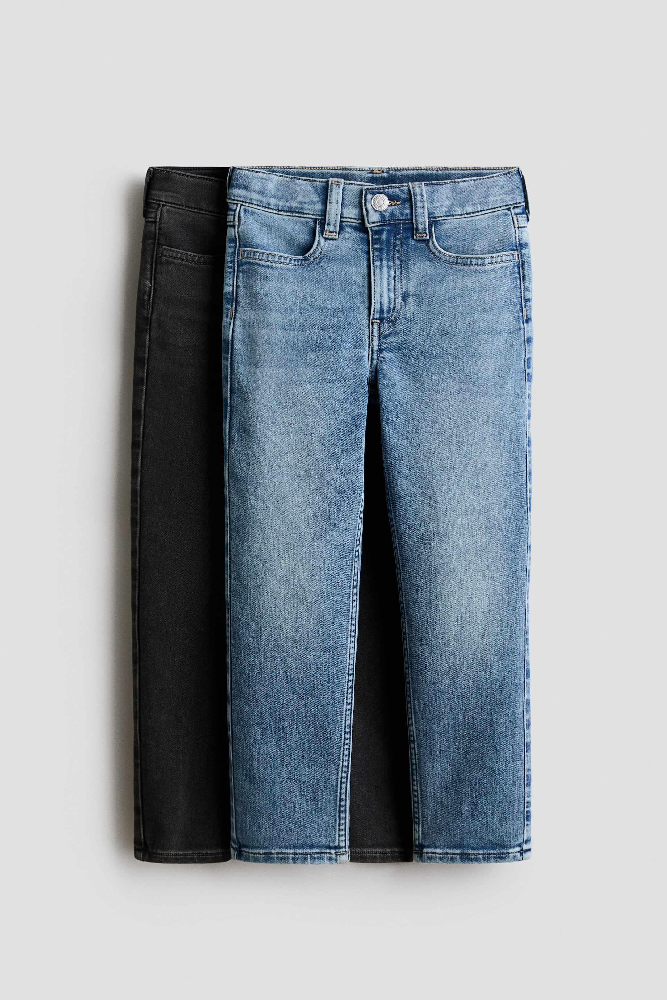 Agrandir l'image: Deux paires de jeans en denim délavé, coupe slim, sont suspendues de face, avec une paire bleu clair superposée directement sur une paire gris foncé. Les deux paires présentent une ceinture classique avec passants de ceinture, une fermeture à bouton métallique, une braguette factice et des poches avant arrondies.