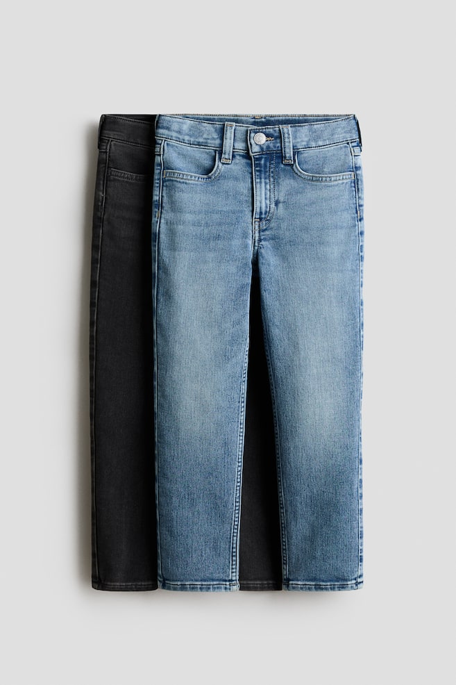 Dois pares de jeans de ganga lavada, de corte slim-fit, estÃ£o pendurados em vista frontal, apresentando um par azul-claro sobreposto diretamente a um par cinzento-escuro. Ambos os pares exibem uma cintura clÃ¡ssica com presilhas para cinto, fecho com botÃ£o metÃ¡lico, braguilha falsa e bolsos frontais curvos. - 1