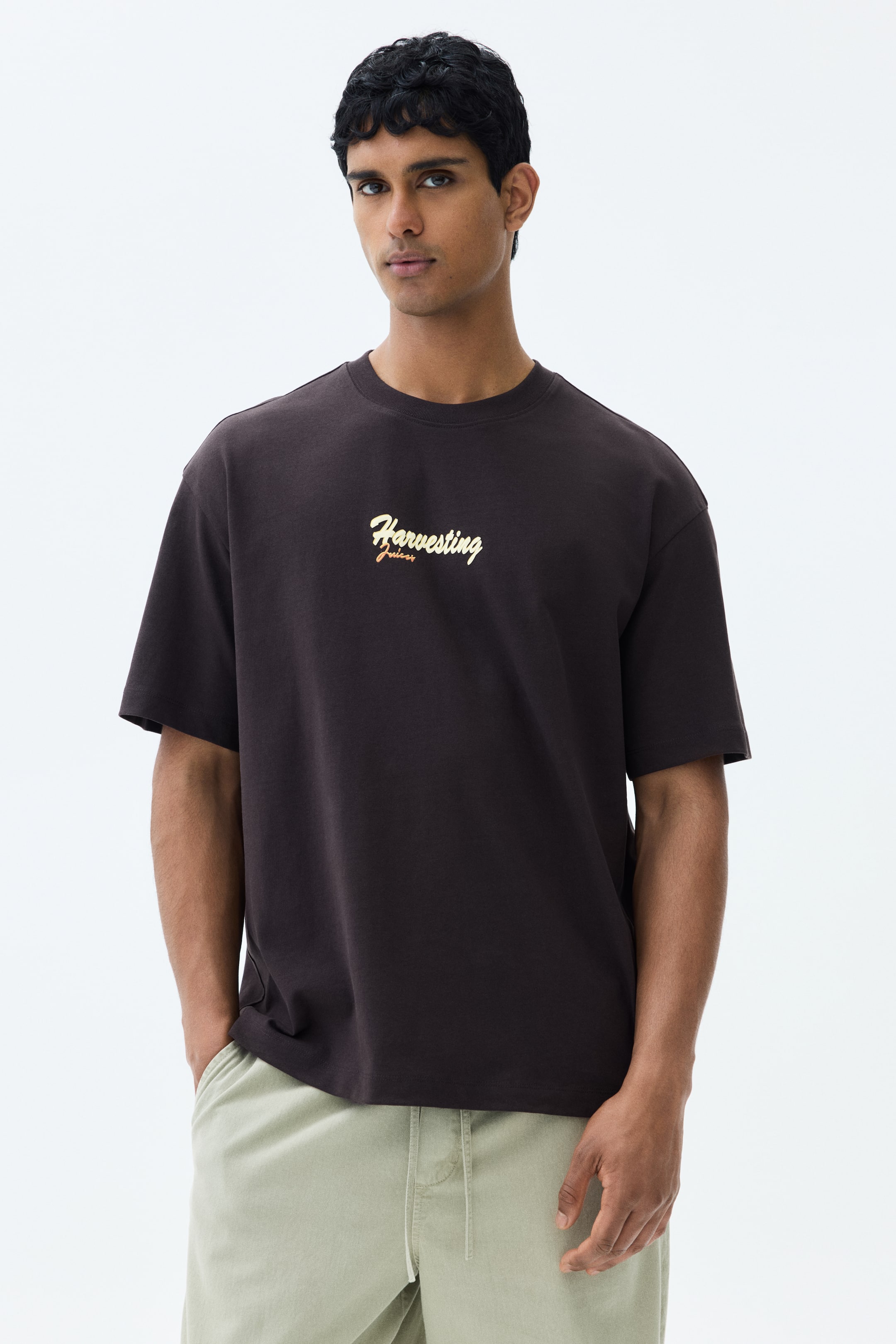 View larger image: The model is donning a dark brown cotton jersey T-shirt, adorned with a golden "Harvesting juices" print. It highlights a round, ribbed neckline, dropped shoulders, a straight hem, and a relaxed fit, with the model's hand casually placed in a trouser pocket.