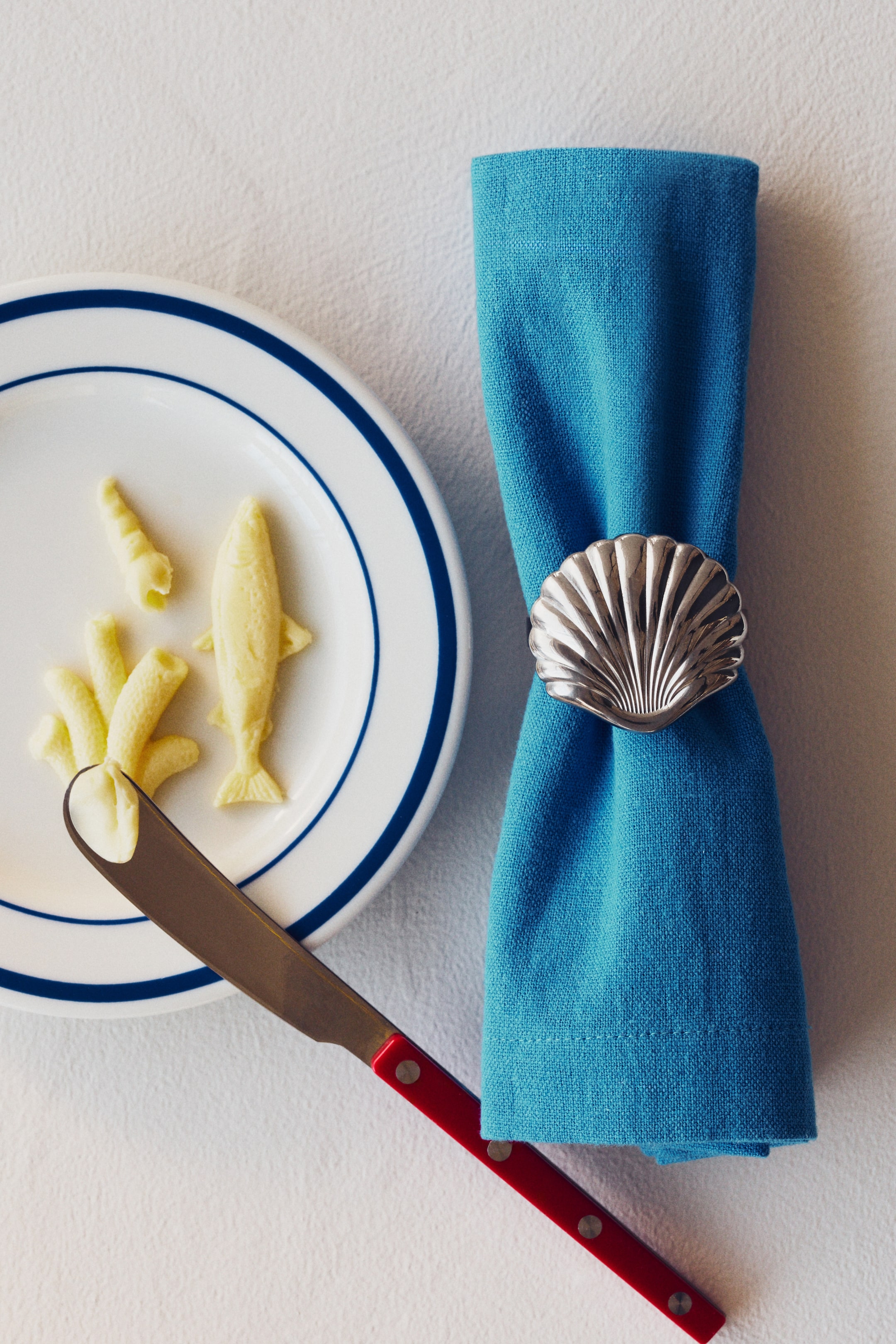 Grotere afbeelding bekijken: Een zilverkleurige servetring in de vorm van een schelp, met duidelijke ribbels op het oppervlak, omringt een gevouwen blauwe stoffen servet en is naar de kijker gericht.