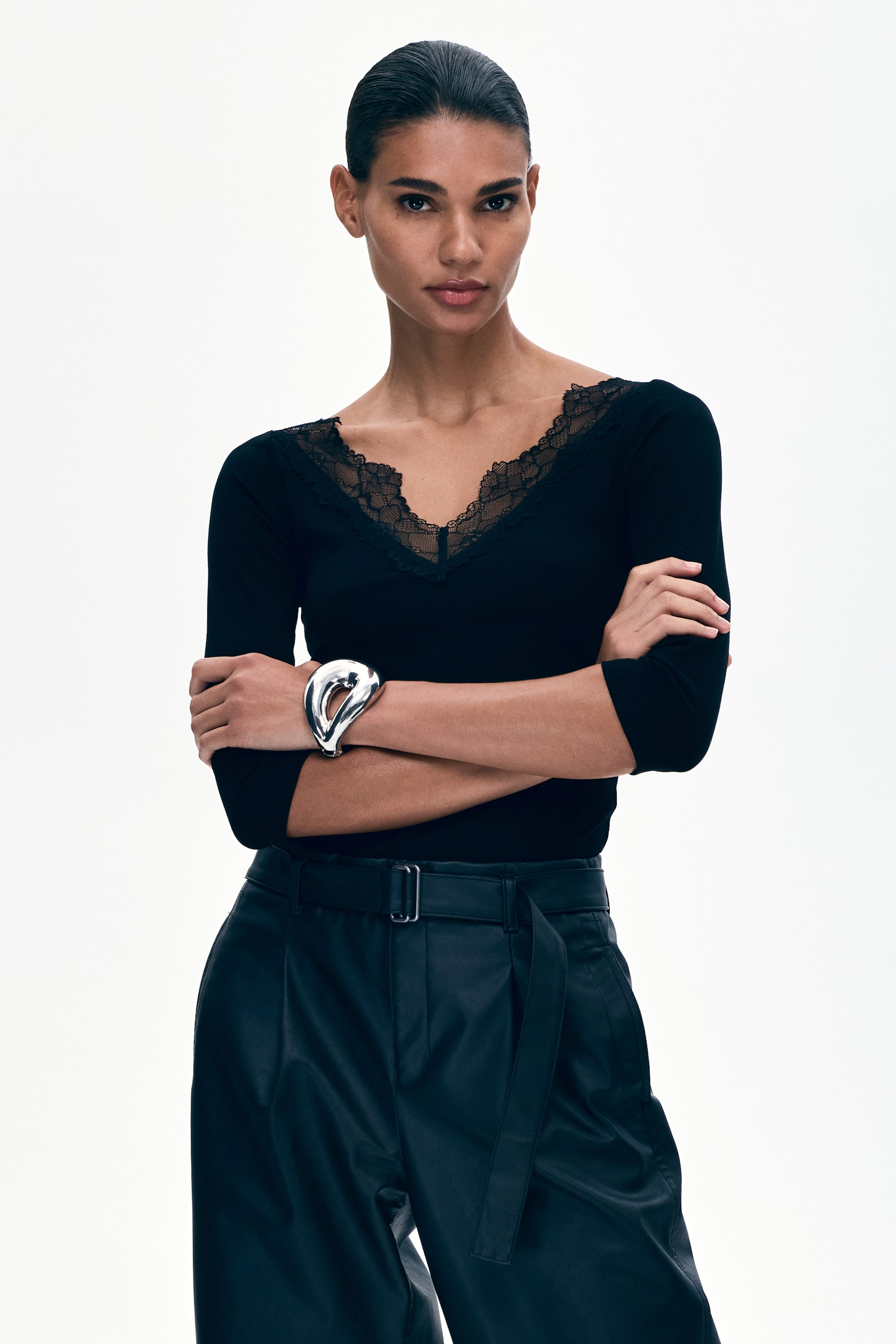 View larger image: The black ribbed knit top boasts a V-neckline adorned with delicate lace trim and three-quarter sleeves. The model is showcasing the top, pairing it with black pleated trousers and a silver cuff bracelet on one wrist.