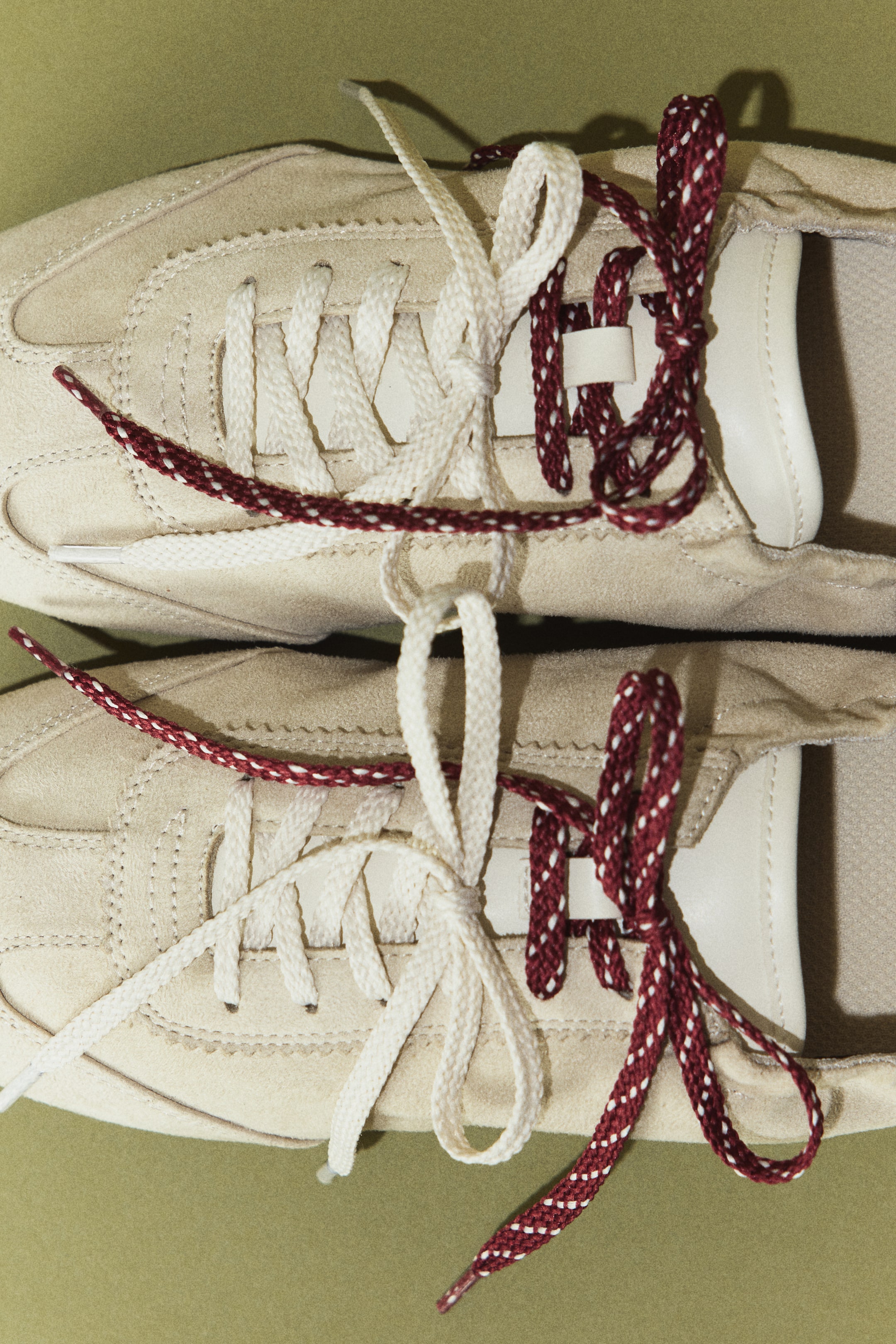Ver imagen más grande: Dos zapatillas de ante beige están colocadas en posición vertical y vistas desde arriba; cada una presenta cordones planos blancos y cordones redondos jaspeados en granate y blanco, entrelazados sobre el empeine.