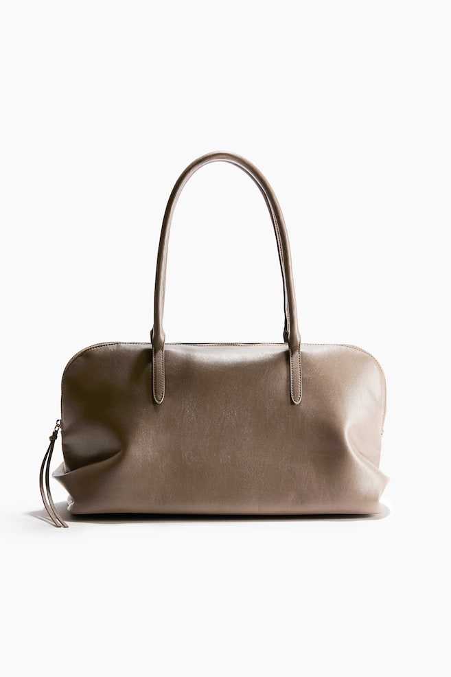 A taupe leather shoulder bag stands upright, facing front to showcase its soft, slouchy rectangular silhouette. It features two long, slender top handles stitched to the front panel and a top zip closure with a long leather tassel pull hanging from the side. - 1