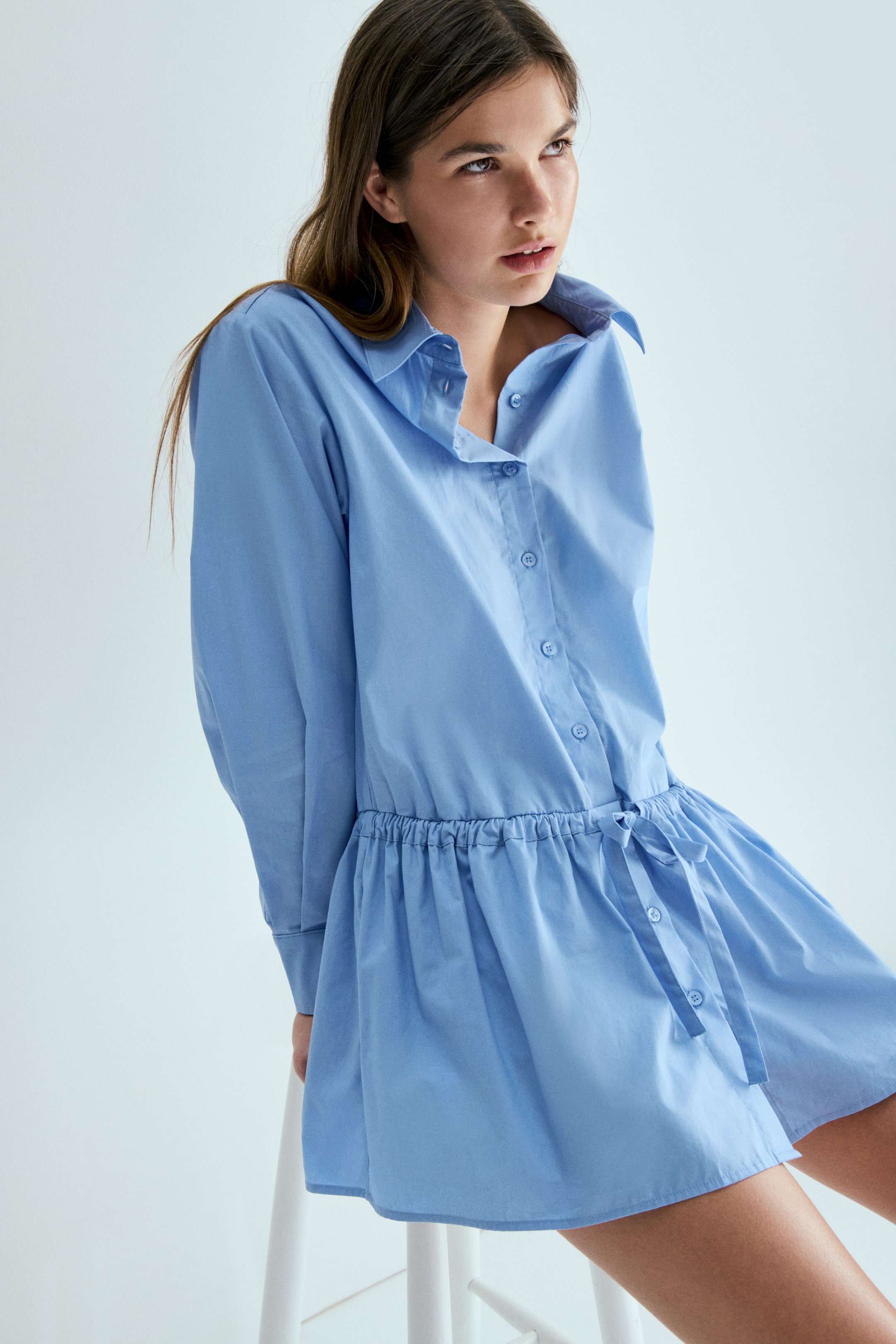 View larger image: A light blue shirt dress featuring a spread collar and button-down placket. It gathers at the waist with an elasticated band and a visible self-tie sash, flowing into a full, gathered skirt.