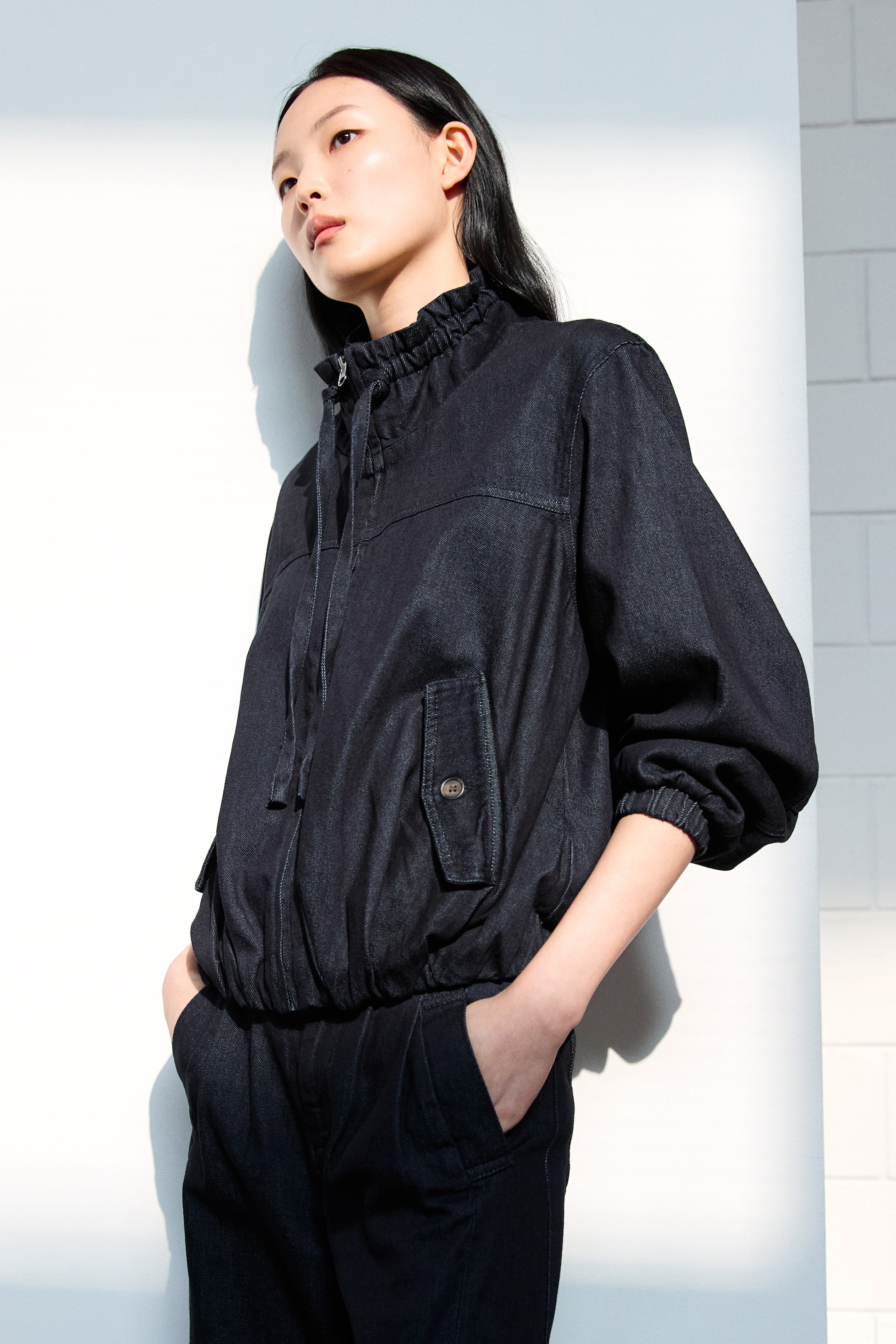 View larger image: A dark indigo denim jacket is featuring a ruched stand-up collar, a front zipper closure with drawstrings, and two buttoned flap pockets. It is showcasing puffed sleeves with elasticated cuffs and an elasticated blouson hem, pairing with matching dark denim pleated pants, viewed from a front-quarter angle.