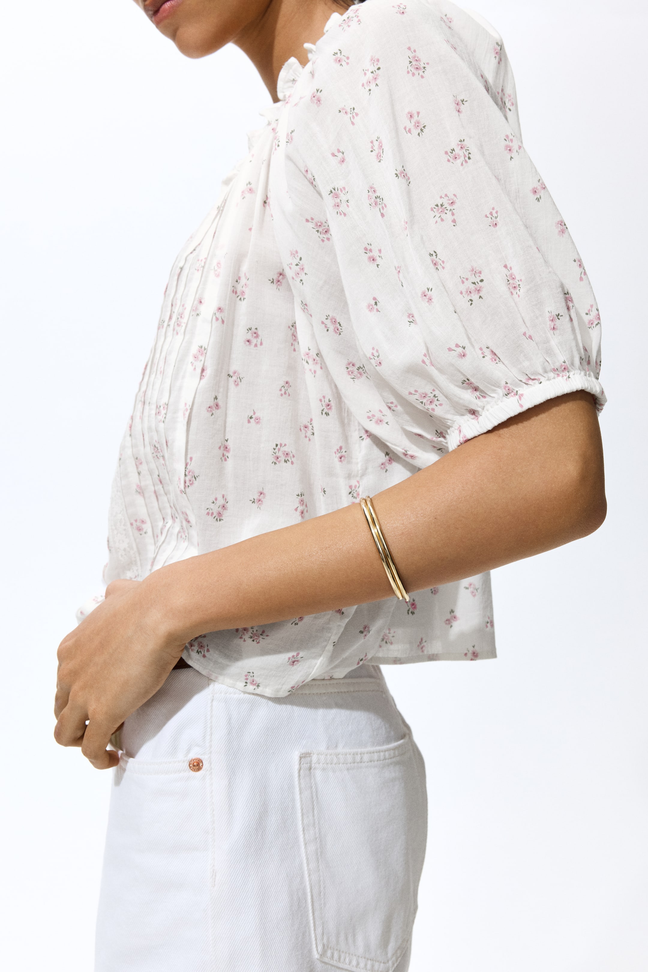 Agrandir l'image: Une blouse blanche avec un imprimé floral rose est portée, vue de côté et de face. Elle présente un col à volants, des plis nervurés verticaux à l'avant, des manches bouffantes avec des poignets élastiqués, et un ourlet court.