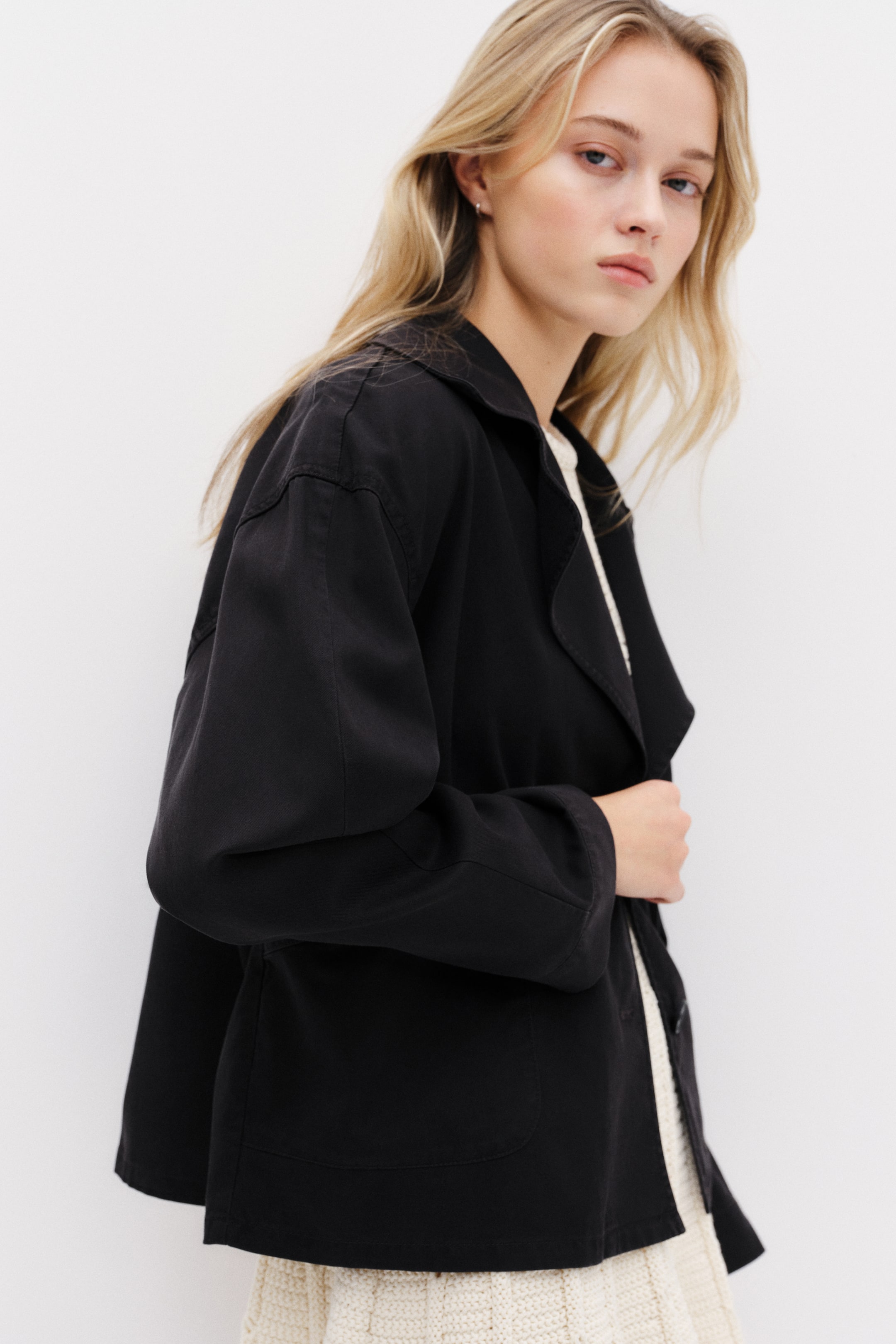 Ingrandisci l'immagine: Una giacca nera presenta ampi revers a lancia e due tasche applicate, con le maniche rilassate che cadono morbidamente. Viene indossata aperta sopra un capo in maglia color crema, mostrando il suo profilo laterale.