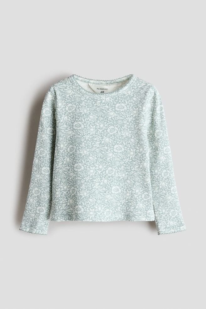Un haut Ã  manches longues ornÃ© dâun imprimÃ© floral vert sauge et blanc sur lâensemble est prÃ©sentÃ© de face, mettant en valeur sa texture en jersey de coton cÃ´telÃ©. Le haut est conÃ§u avec un col rond et des poignets, tous deux rehaussÃ©s dâune dÃ©licate finition picot. - 1