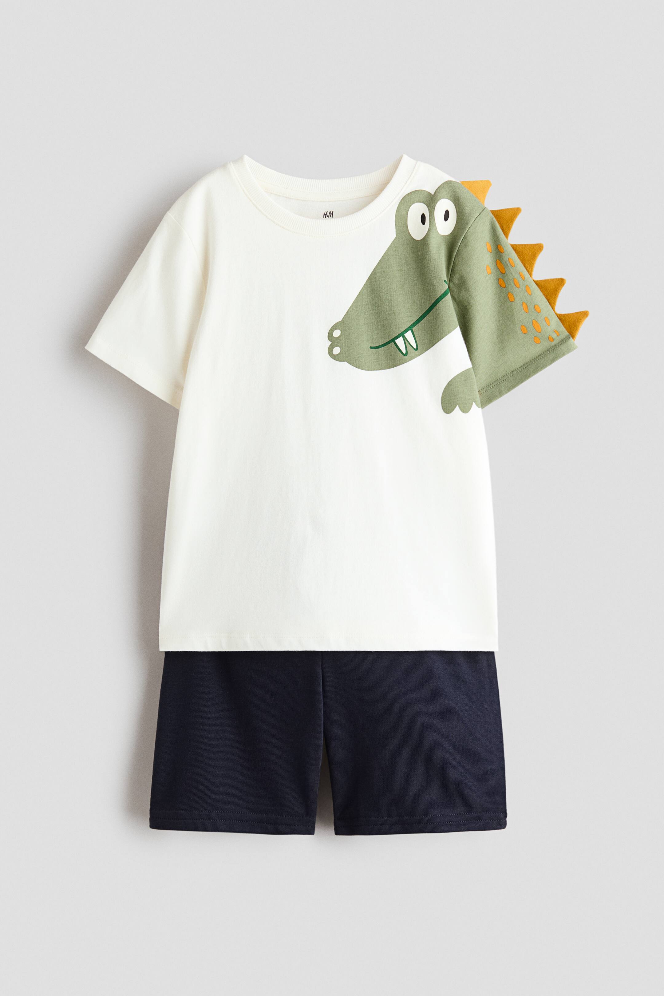 查看大圖: 一套童裝兩件式服裝正展示正面視圖,米白色T恤配搭海軍藍短褲。T恤採用圓領設計,左側印有綠色鱷魚圖案,袖縫位置延伸出橙色布料鋸齒裝飾。配襯的針織短褲垂掛於下方,設計簡潔,為直筒褲管款式。